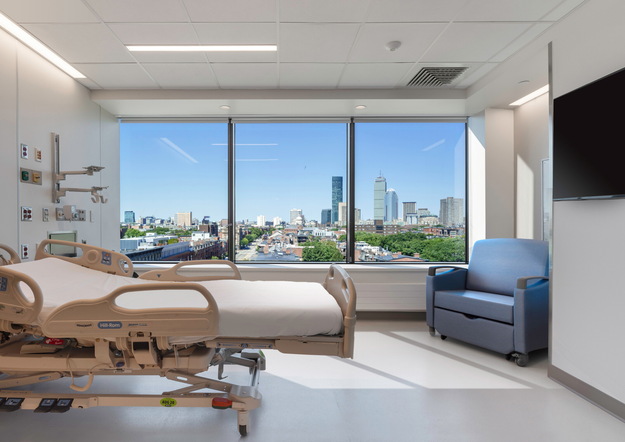 Modern hospital room with a bed, medical equipment, and a seating area by a large window