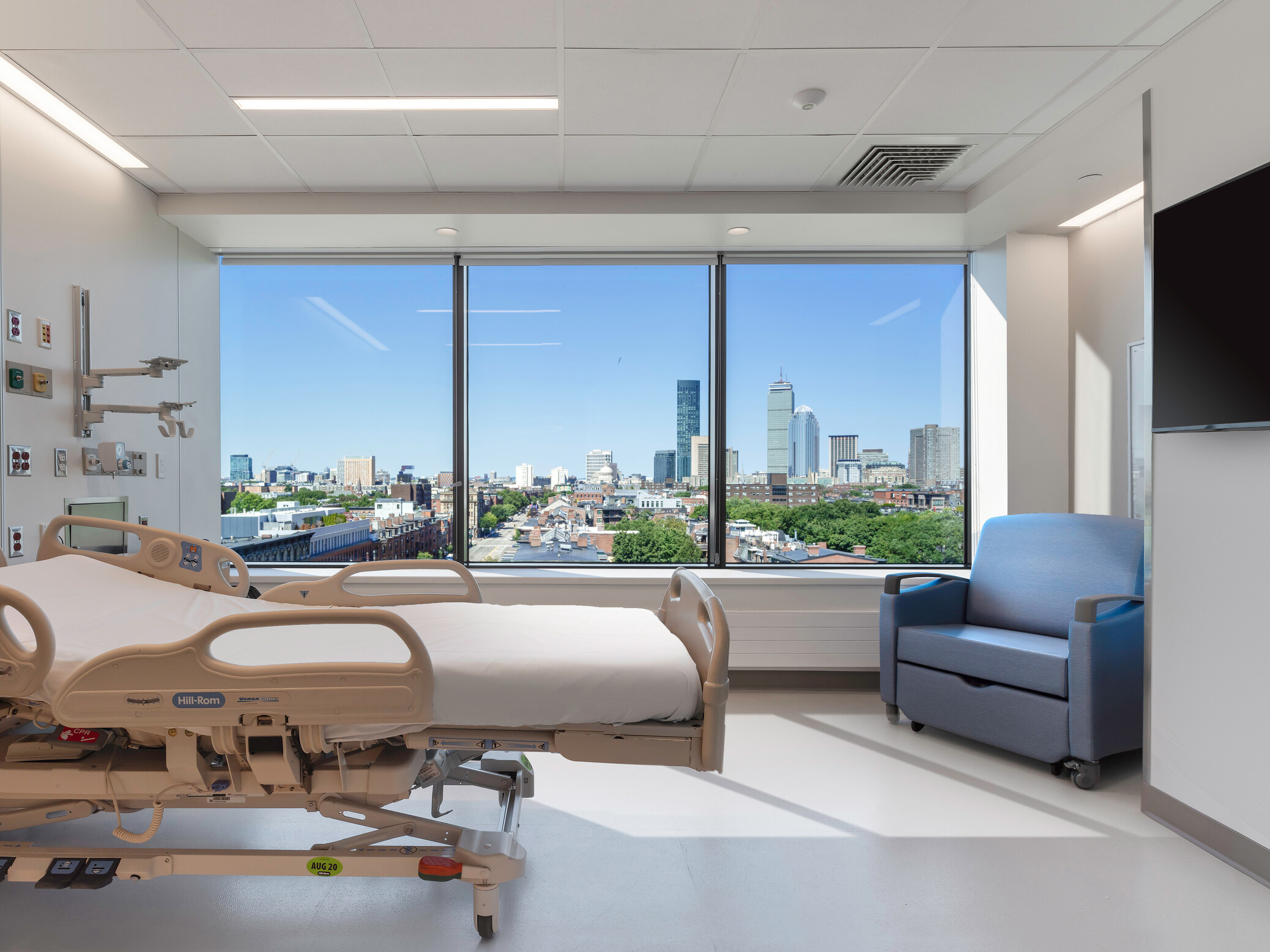 Modern hospital room with a bed, medical equipment, and a seating area by a large window