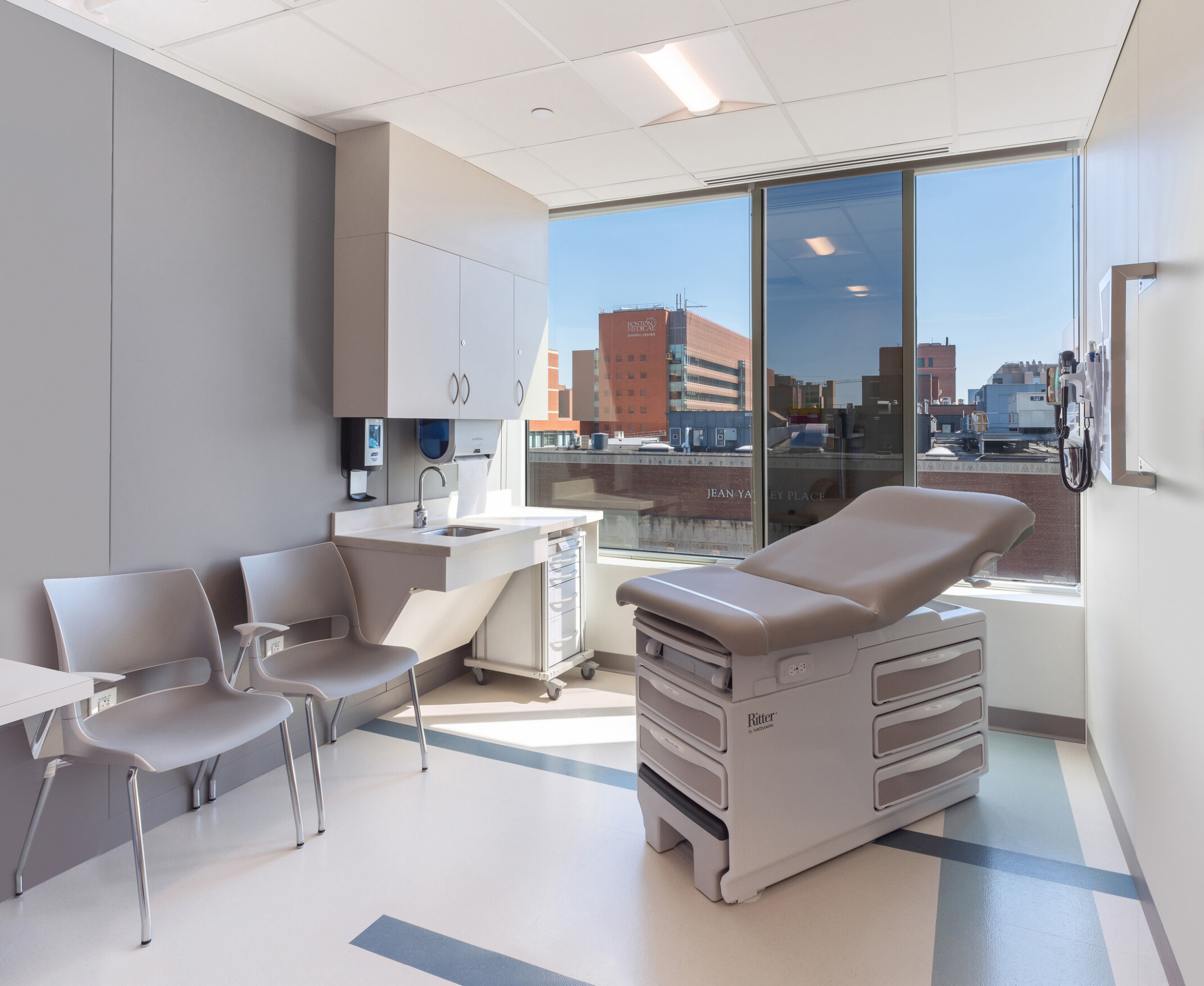 Modern hospital room with an exam table, medical equipment, and a seating area by a large window