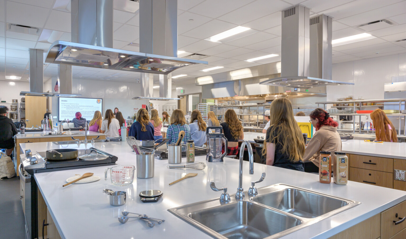 kitchen area for class-style learning pictured from the back; children at each station learning from an instructor in the front of the classroom; kitchen utensils clutter the counterspace in the foreground
