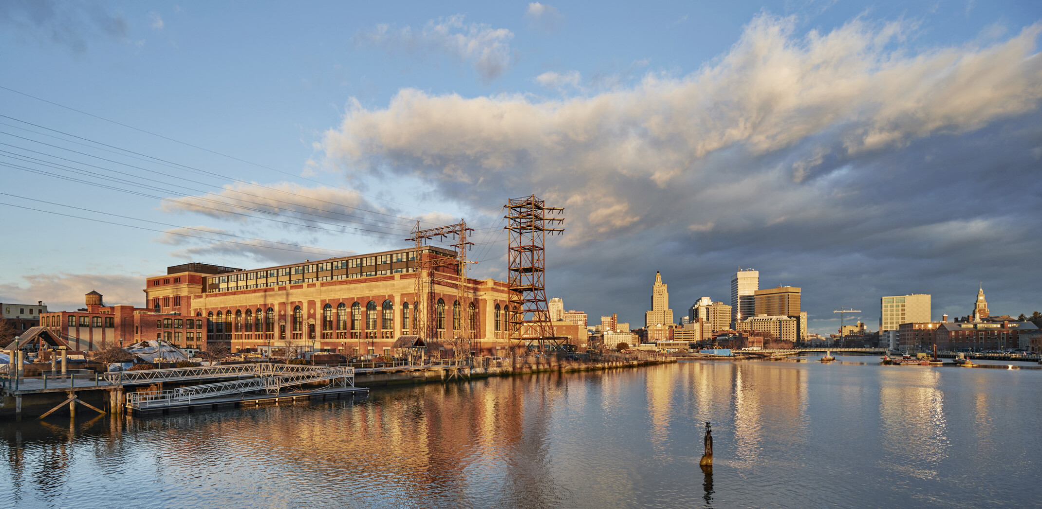 River view of South Street Landing in Providence, RI; adaptive reuse of an old power station into medical training facility; large building with older facade and power lines surrounding River view of South Street Landing in Providence, RI; adaptive reuse of an old power station into medical training facility; large building with older facade and power lines surrounding