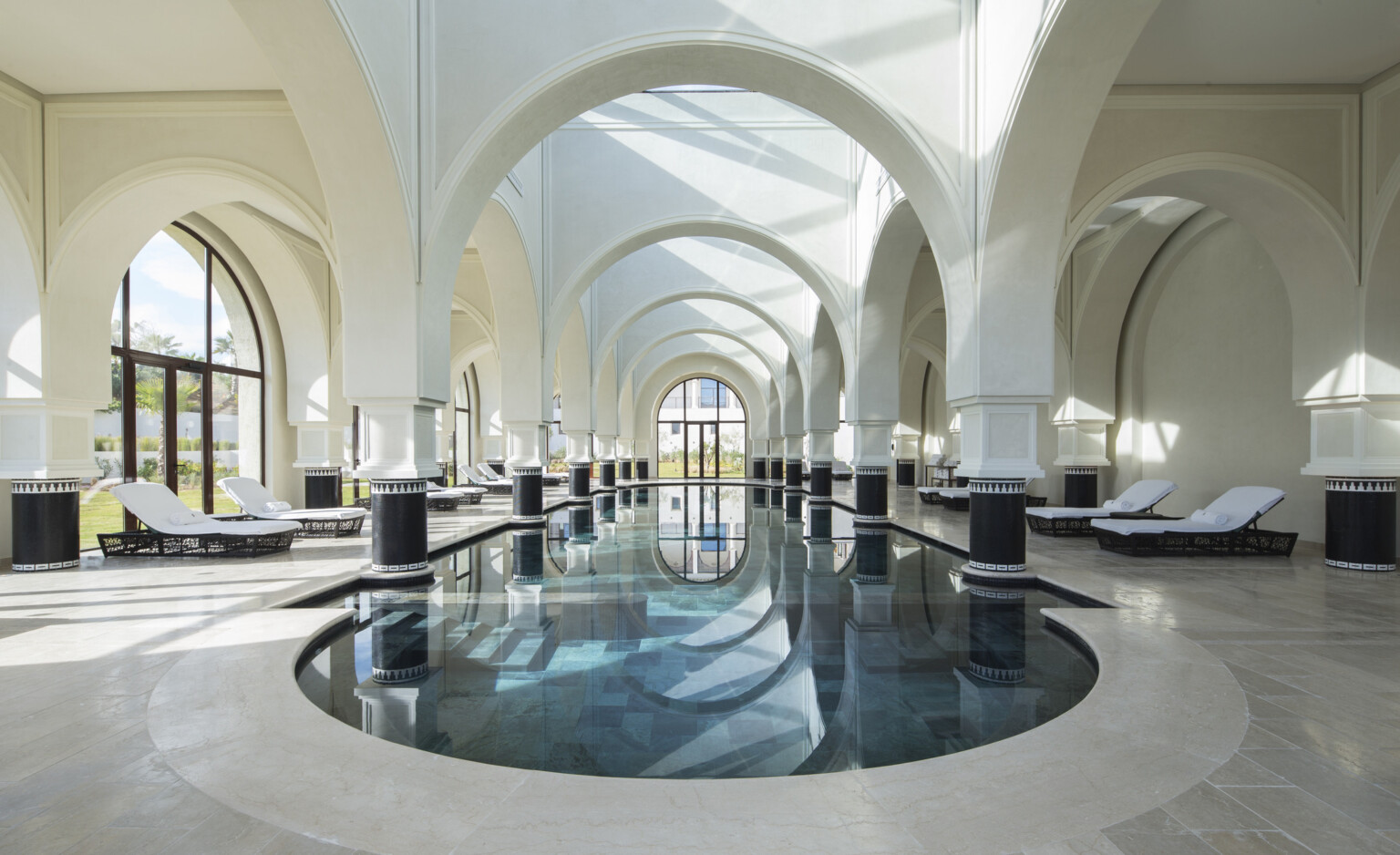 Luxurious indoor pool lined with lounge chairs. High vaulted arches throughout and columns with black bases in the pool