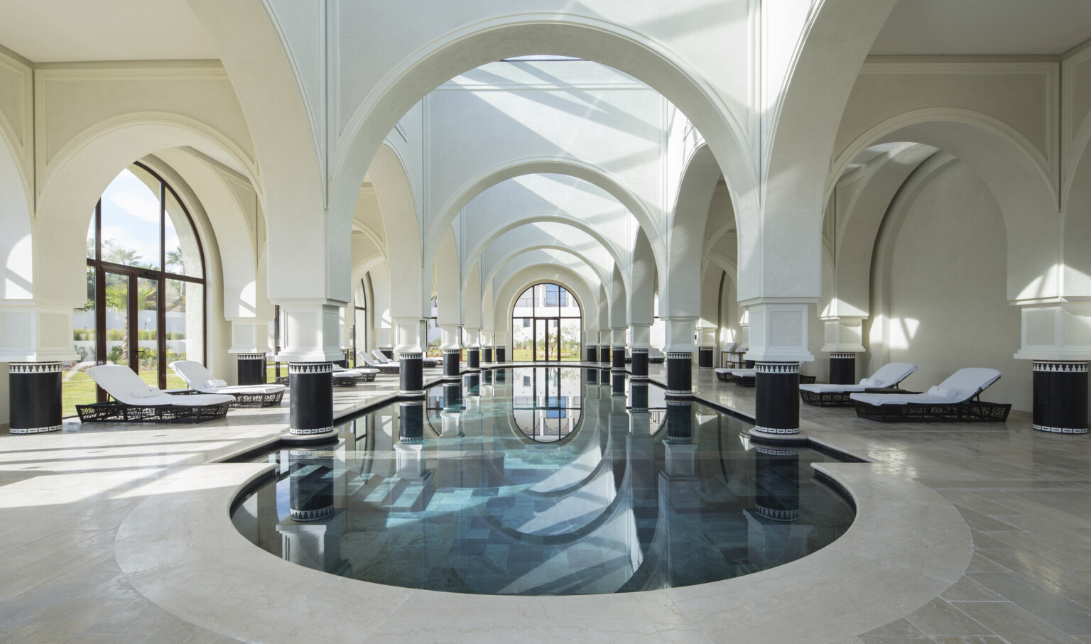 Luxurious indoor pool lined with lounge chairs. High vaulted arches throughout and columns with black bases in the pool