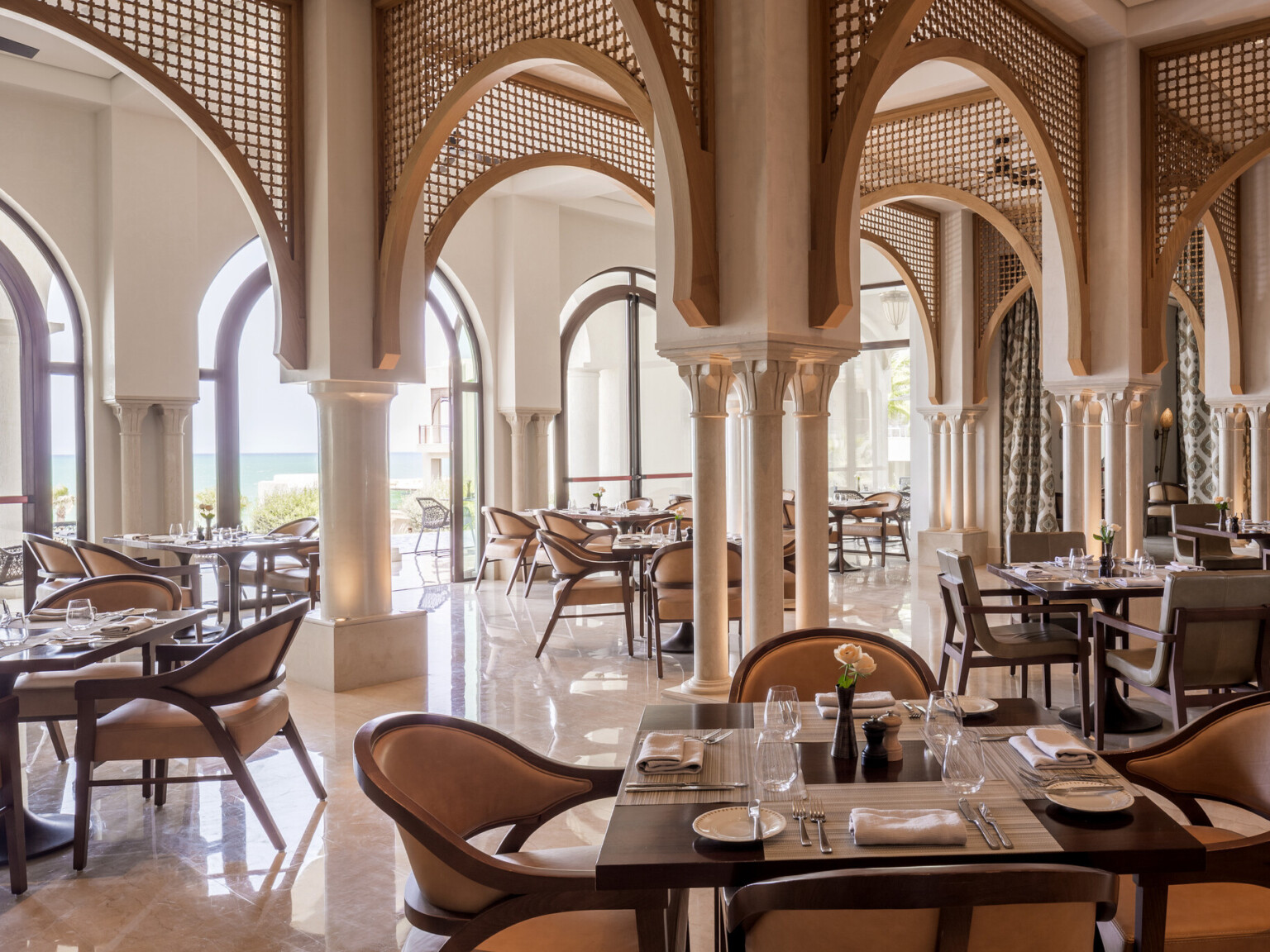 Luxurious restaurant filled with tables and chairs with arched windows overlooking the ocean. Multiple columns throughout with ornate latticework