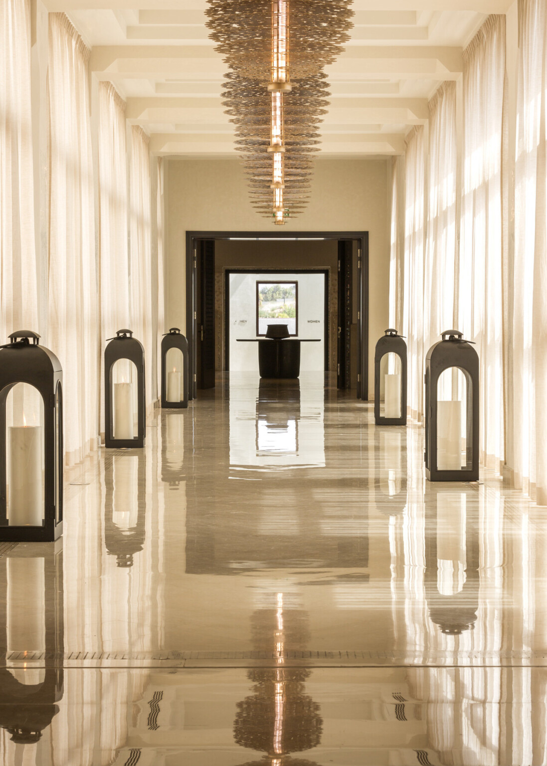 A long hallway lined with large black-framed lanterns, sheer curtains, and a modern chandelier
