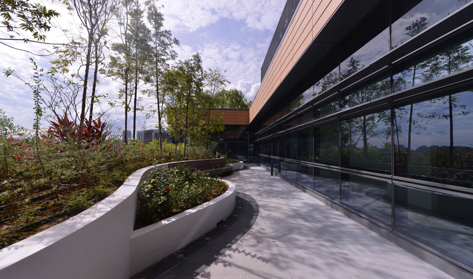 Modern building with large glass windows, a curved pathway, and a landscaped garden enclosed by a white wall under a partly cloudy sky.