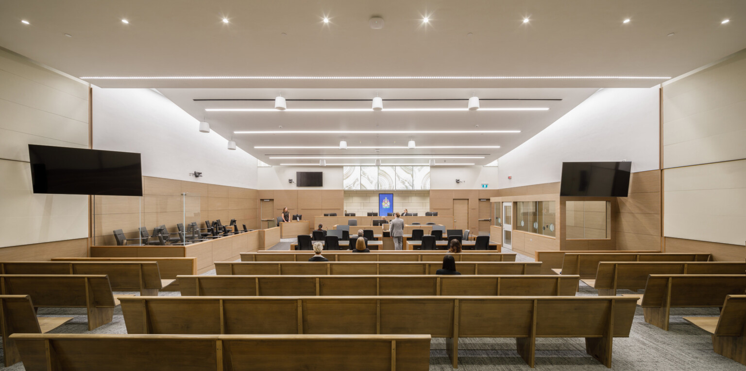 wide view looking through a large courtroom to the judge's bench at the center of the front wall. Rows of bench seating flank two aisles, digital screens on the side walls.