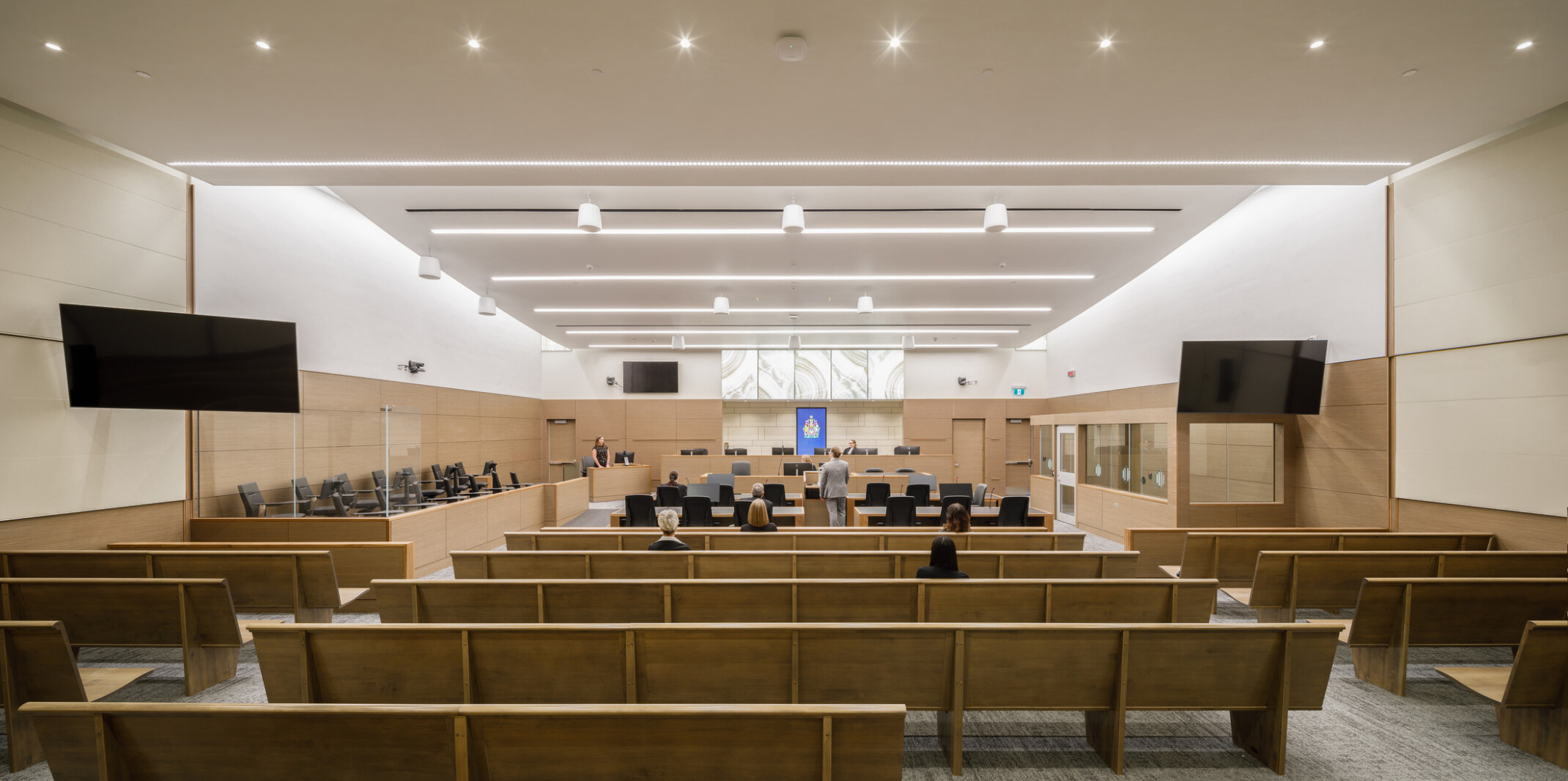 wide view looking through a large courtroom to the judge's bench at the center of the front wall. Rows of bench seating flank two aisles, digital screens on the side walls.
