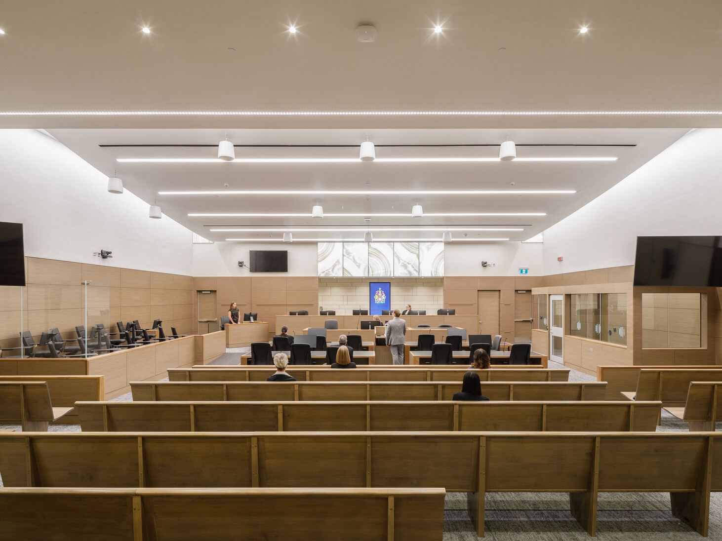 wide view looking through a large courtroom to the judge's bench at the center of the front wall. Rows of bench seating flank two aisles, digital screens on the side walls.