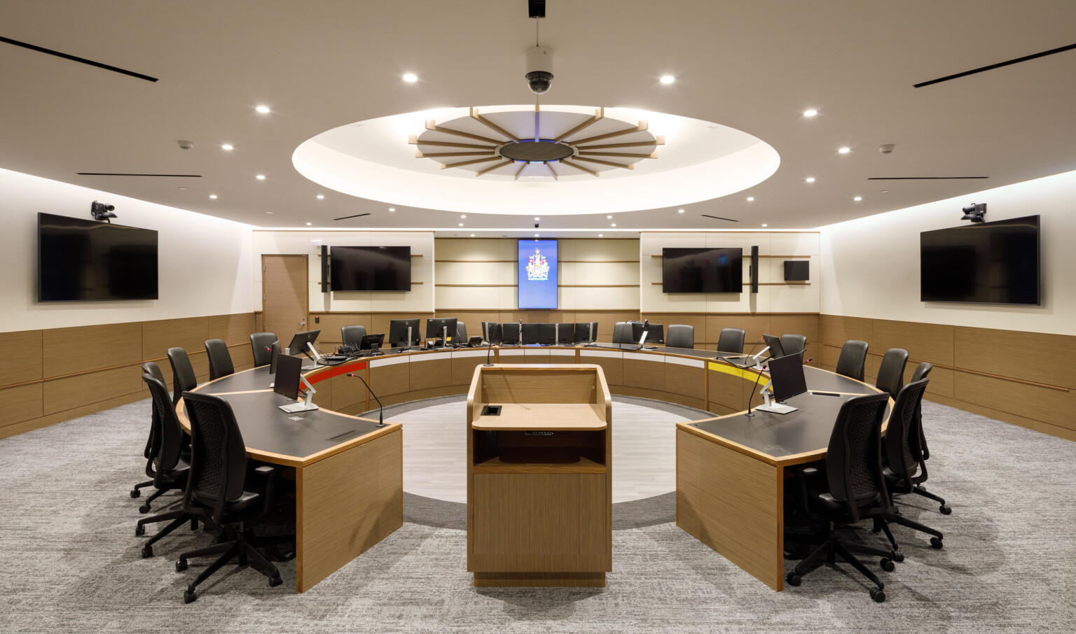 View of indigenous courtroom with a semi-circular table for 20-plus people. Digital screens and cameras mounted on the walls and a recessed circular light fixture above the table.