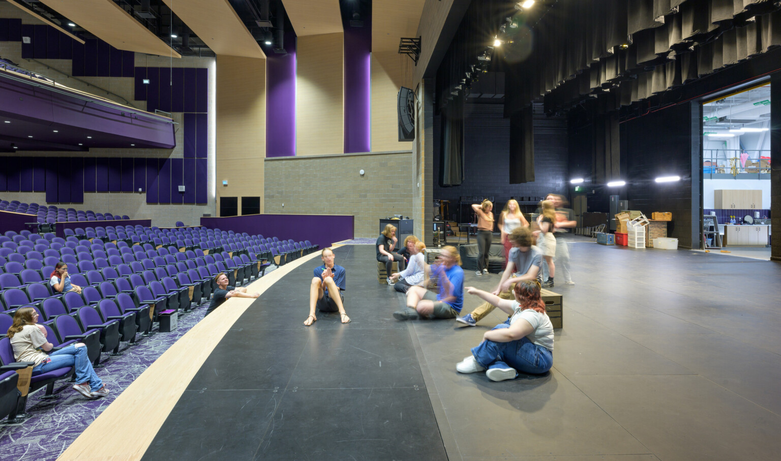 Auditorium pictured from stage left with children conversing and moving around the stage; blue seats scattered with students face the fully lit stage