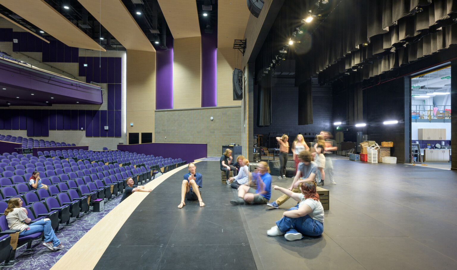 Auditorium pictured from stage left with children conversing and moving around the stage; blue seats scattered with students face the fully lit stage