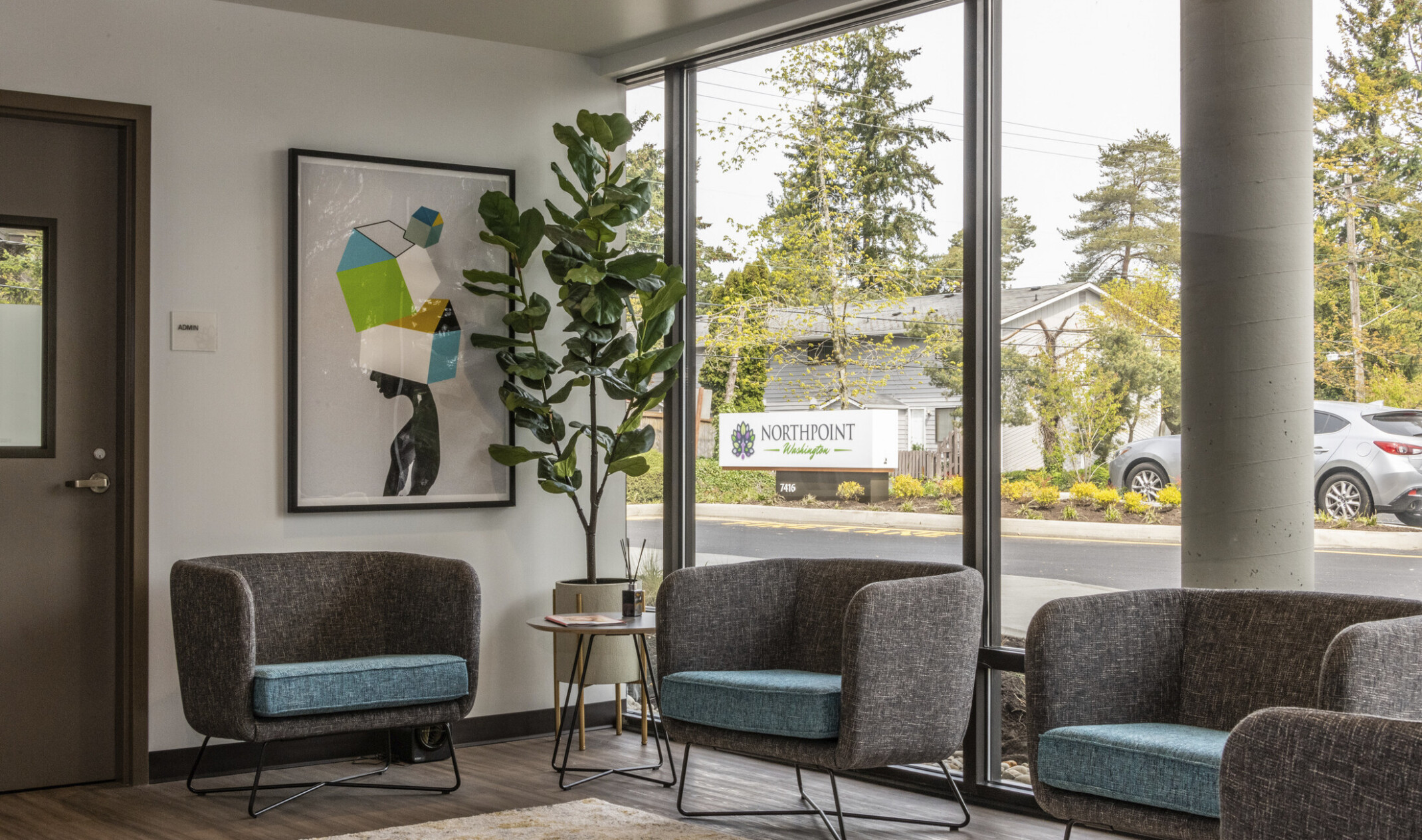 a seating area with gray upholstered bucket seating and blue seat cushions in front of floor to ceiling glass & abstract art