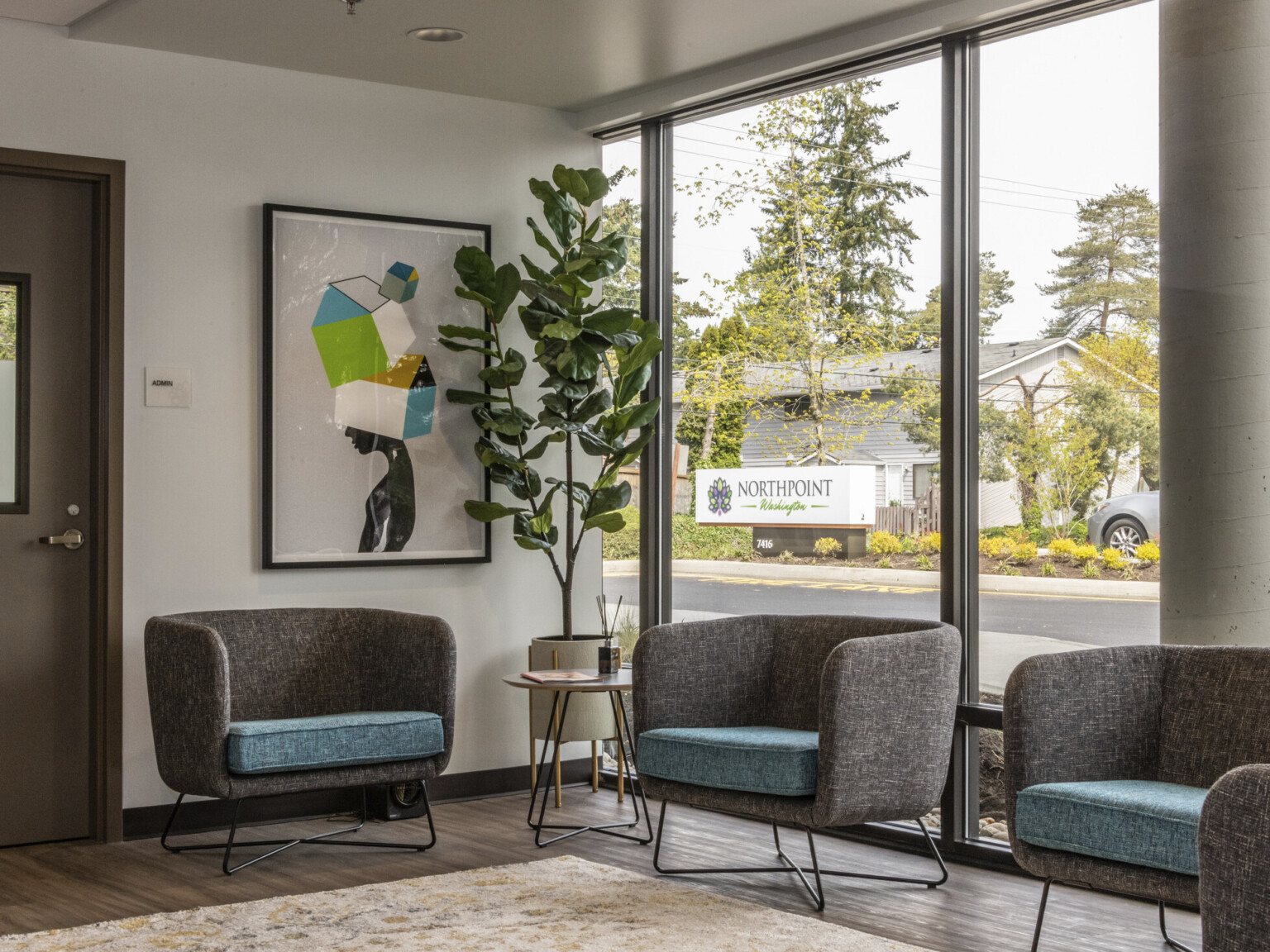 a seating area with gray upholstered bucket seating and blue seat cushions in front of floor to ceiling glass & abstract art