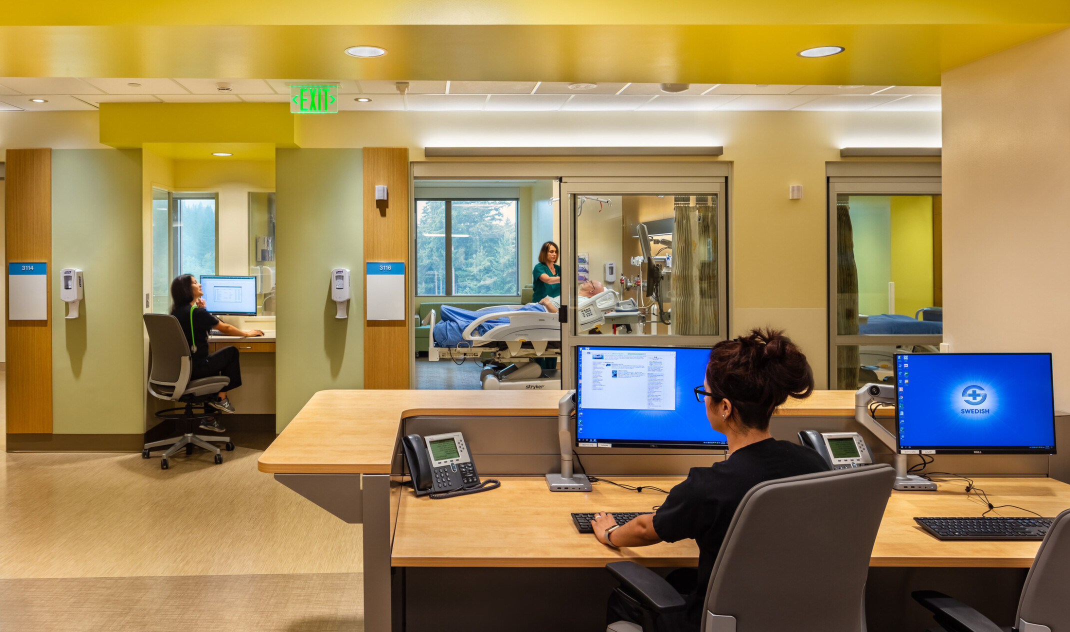 view of a hospital hallway and bed area from behind the nurses station; people working at computers