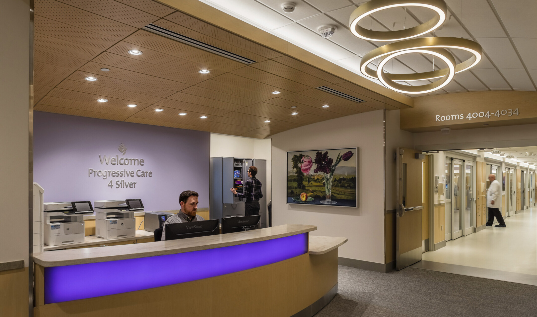 Reception desk with illuminated blue panel on front under modern round chandeliers in hallway. Office behind blue accent wall
