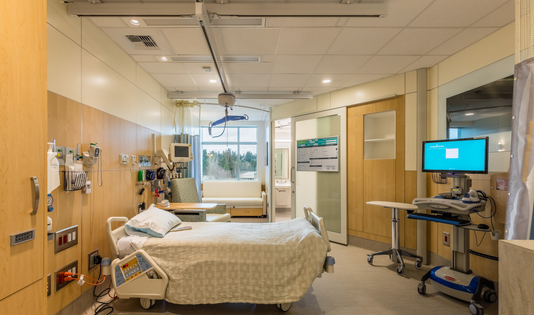 hospital room; single bed facing mobile computer stand; warm wood and warm white accented elements