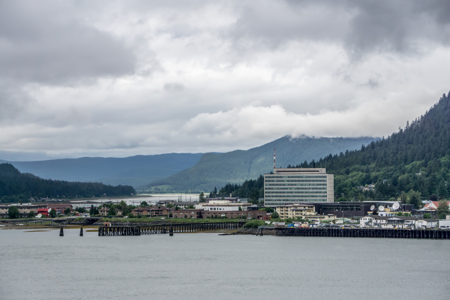 Scenic view of the Juneau Federal Building and U.S. Courthouse, with the Gastineau Channel in the foreground and fir-covered coastal hills in the background. Sky is gray with low clouds Scenic view of the Juneau Federal Building and U.S. Courthouse, with the Gastineau Channel in the foreground and fir-covered coastal hills in the background. Sky is gray with low clouds