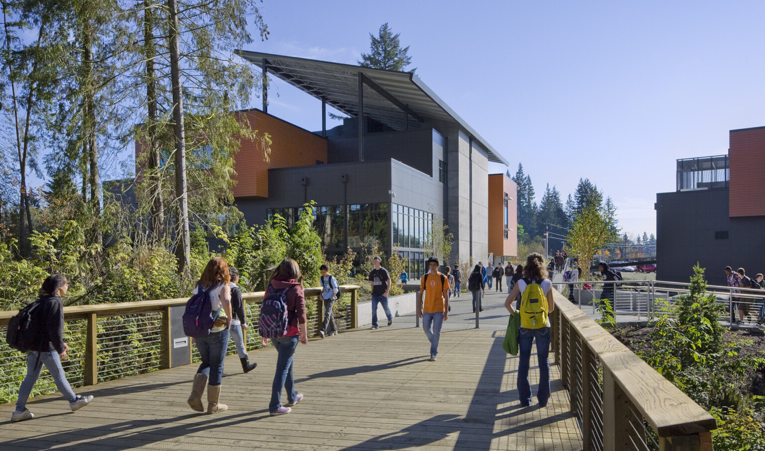 Wood bridge over trees looking to grey 3 story building with orange panel accents and large roof panel angled across top