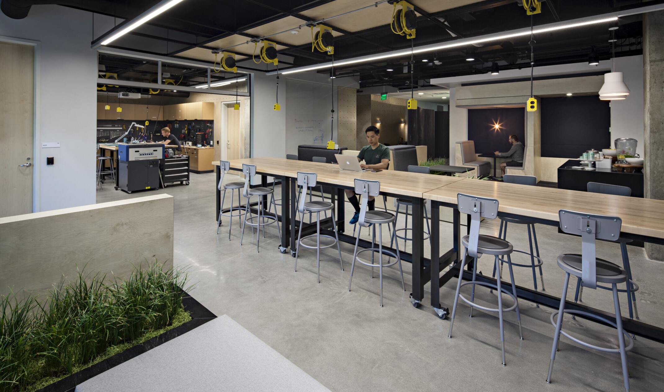 Office lounge with industrial seating surrounding a light wood table, yellow and black electrical outlets suspended from the ceiling and a workshop in the background with cabinets lining the walls and a high top table in the center of the room