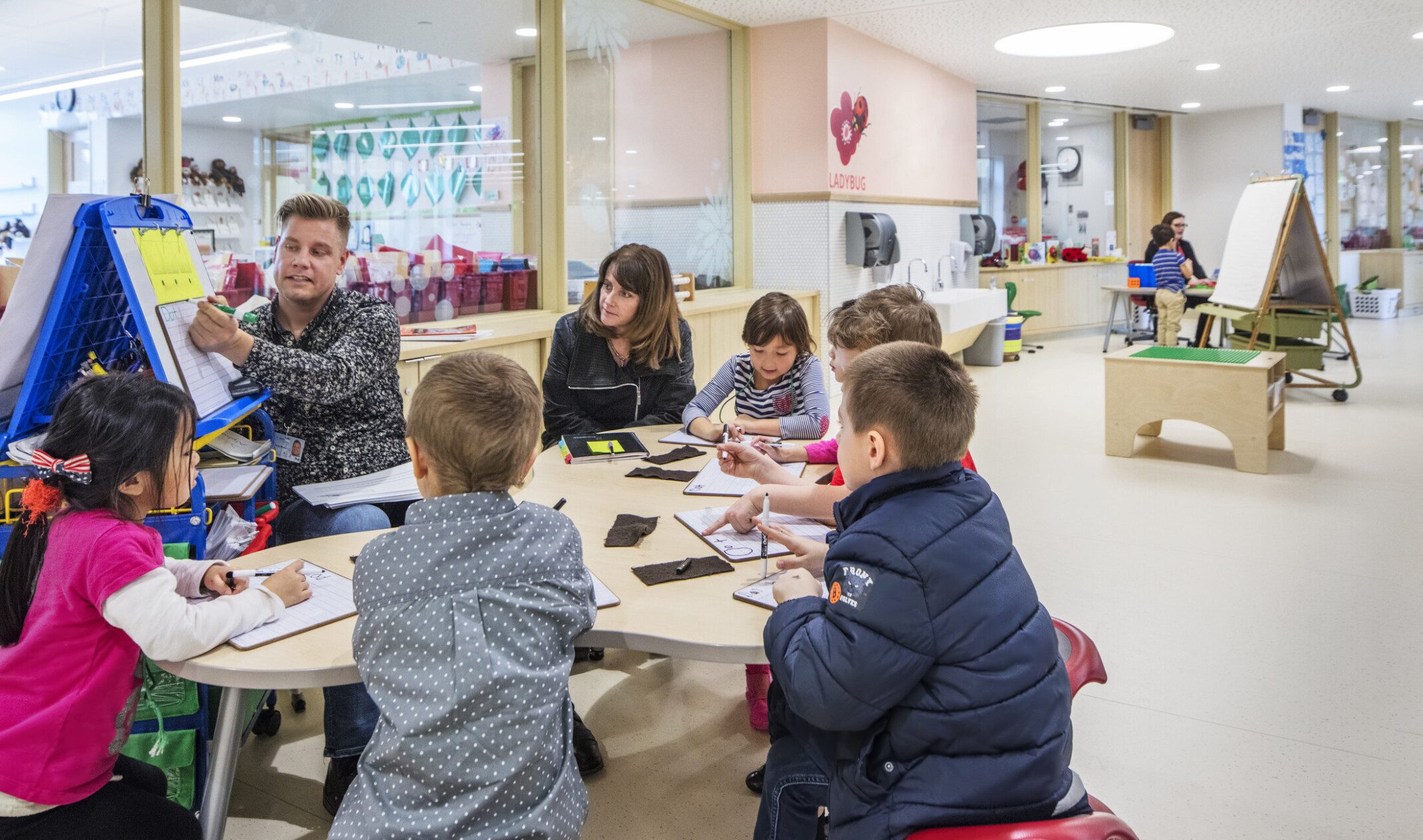 Group of children gathered around a table listening to a teacher teach off of a whiteboard