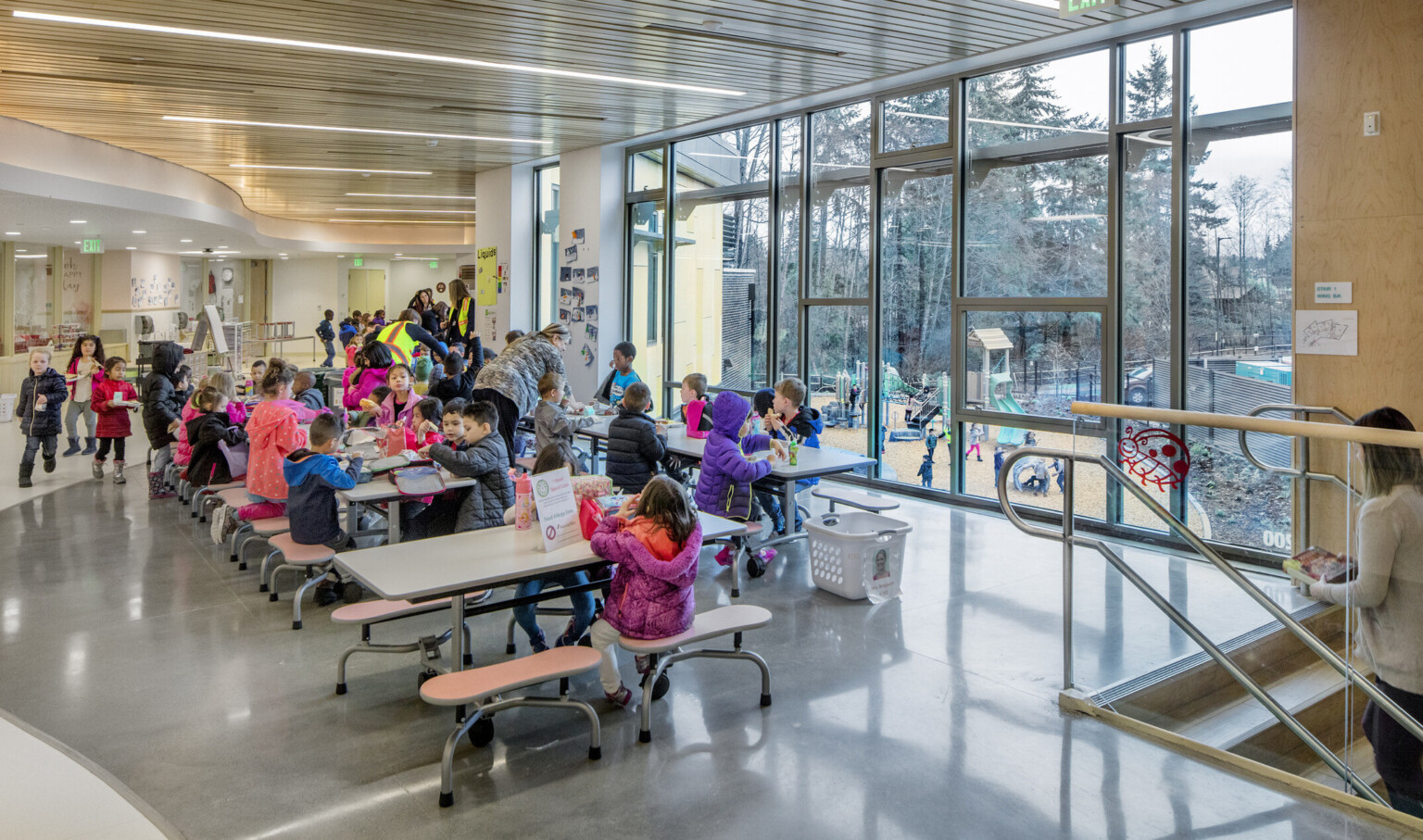 A lunch room with floor to ceiling windows with views of the playground. Above, a curved wood ceiling detail