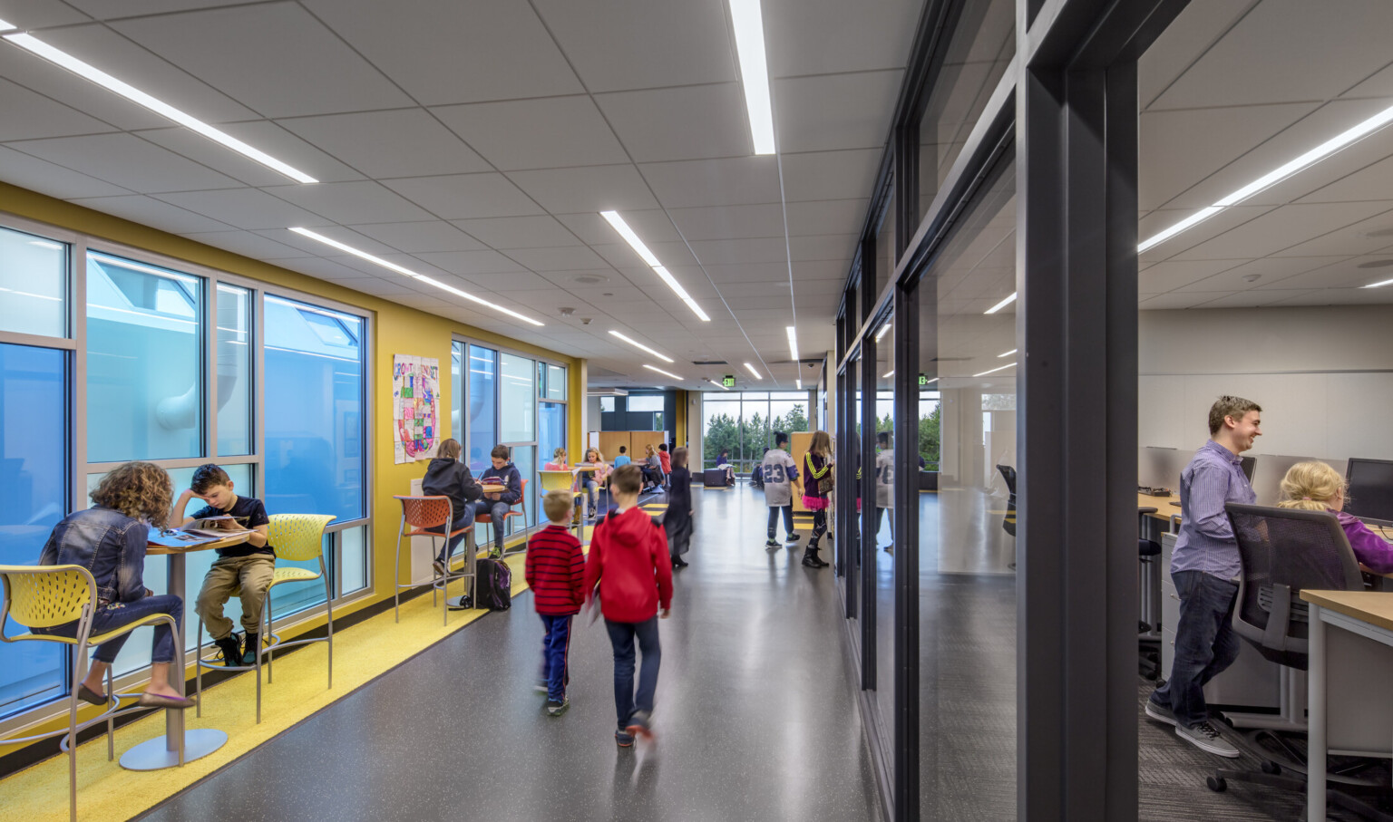 Interior hallway with yellow wall, left, with blue translucent windows. Glass walls into classrooms, right. High top tables