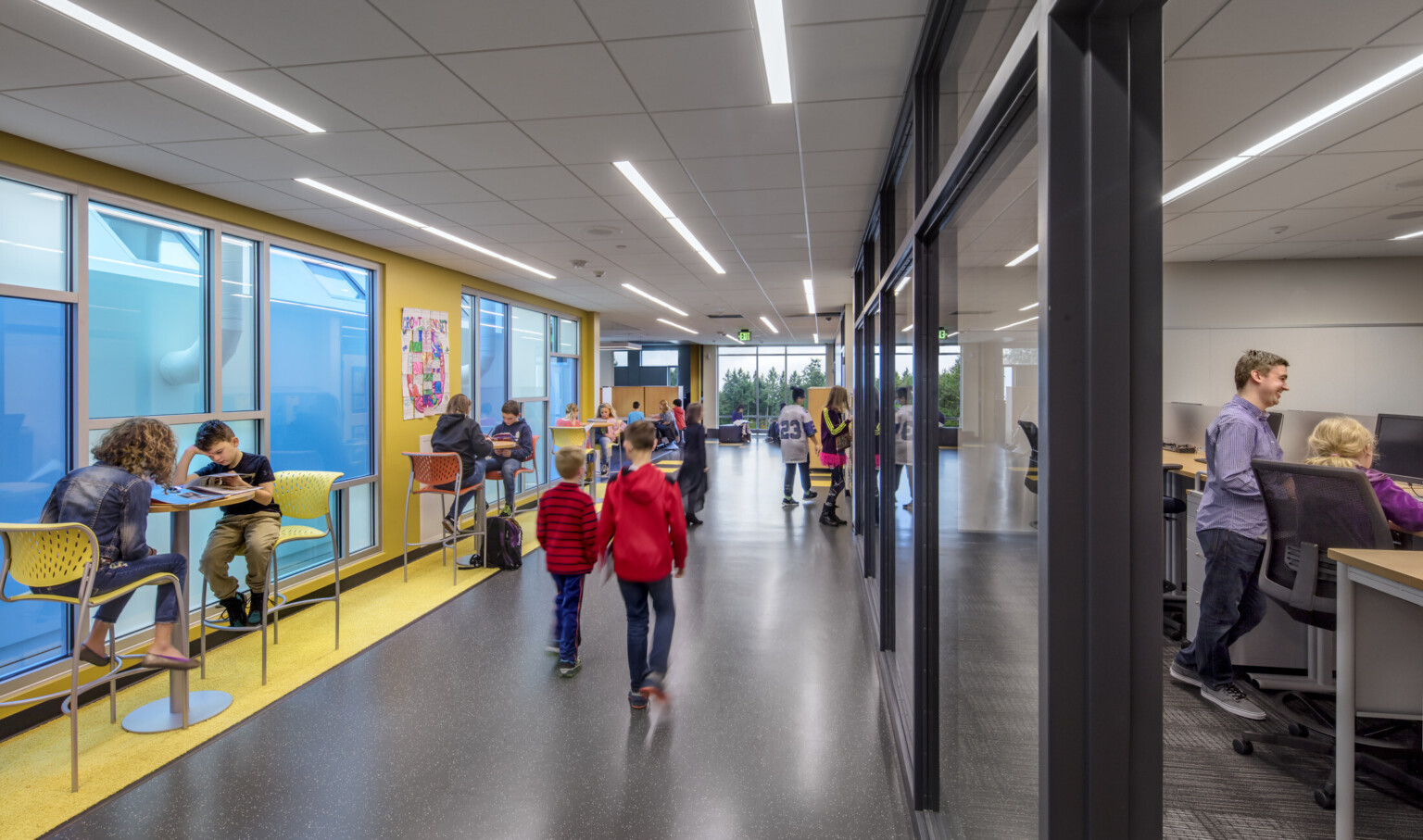 Interior hallway with yellow wall, left, with blue translucent windows. Glass walls into classrooms, right. High top tables