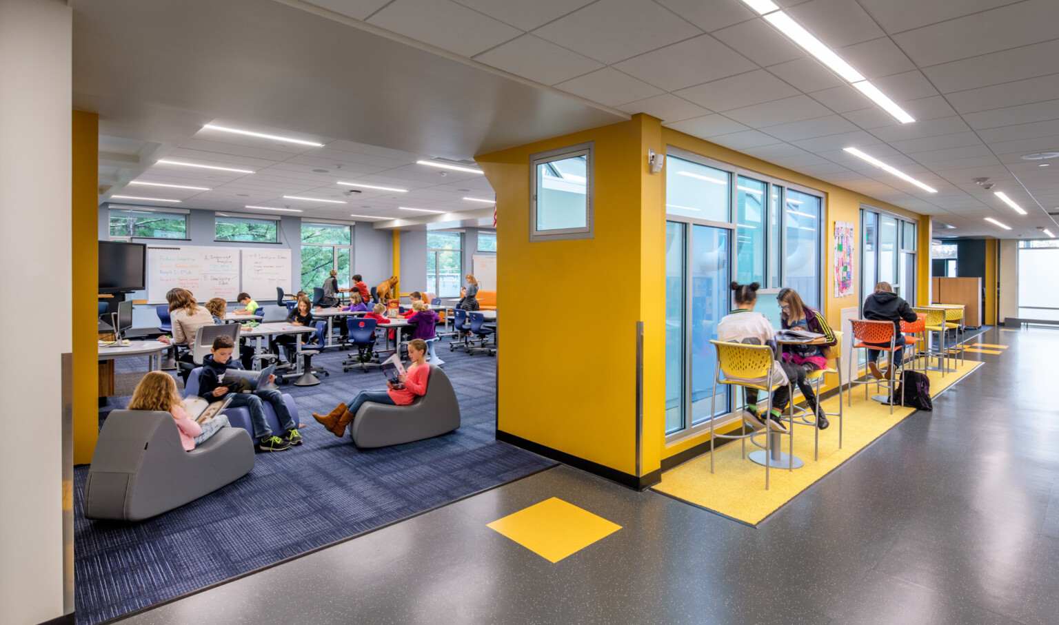 Hallway with high top tables and chairs separated from flexible and ergonomic seating by yellow wall with sheer blue windows