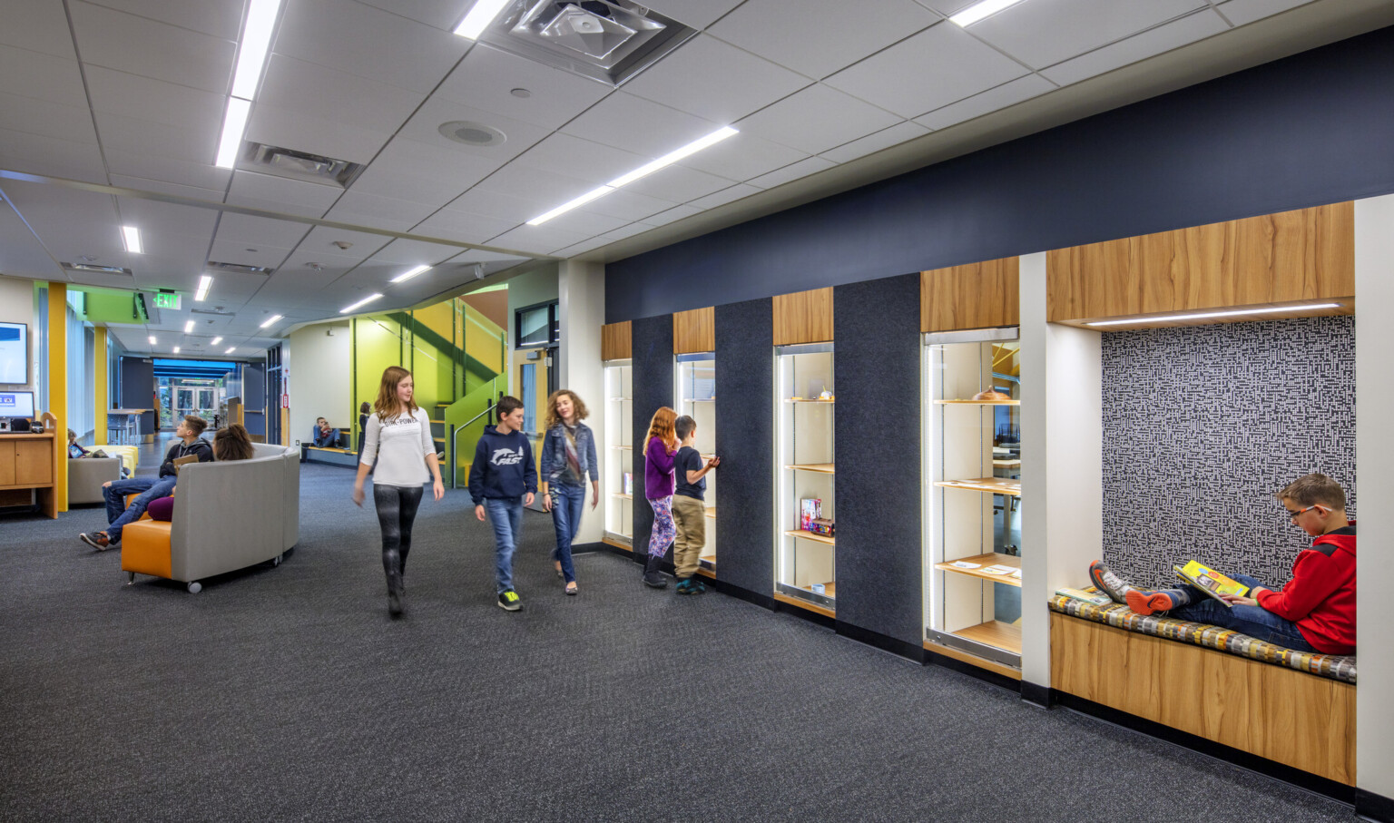 Hallway with blue wall with wood accents, recessed seating and display shelves. Hall leads to seating and green stairwell
