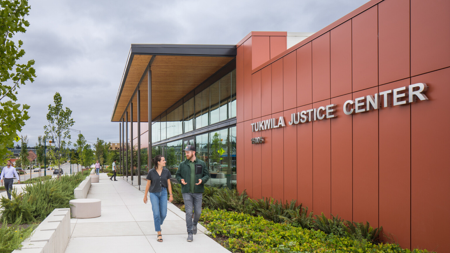 City of Tukwila Justice Center on overcast day, with landscaping in the foreground, trees, single-story double-height glass façade building, large signage, people strolling on front entrance walkway