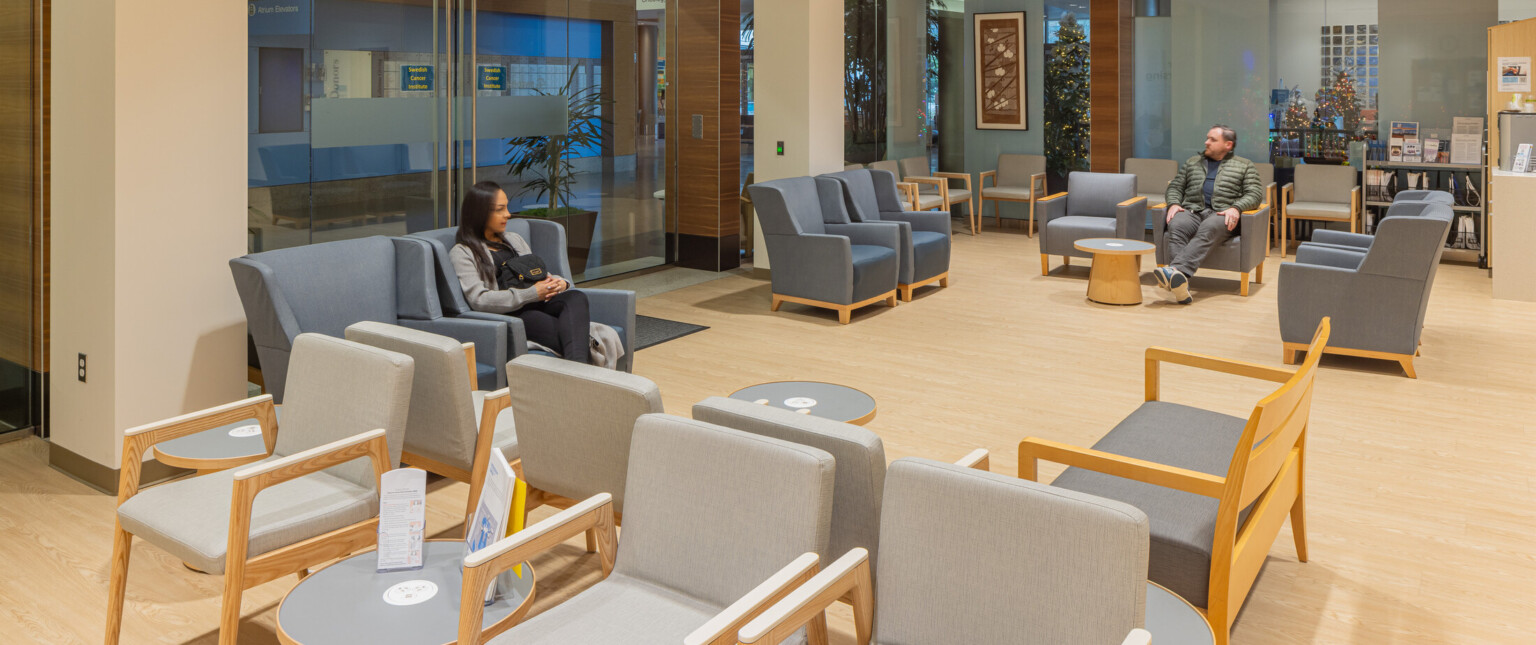 Medical waiting room with light wood floors, grey fabrics in a row in front of blue chairs arranged in a square around a table