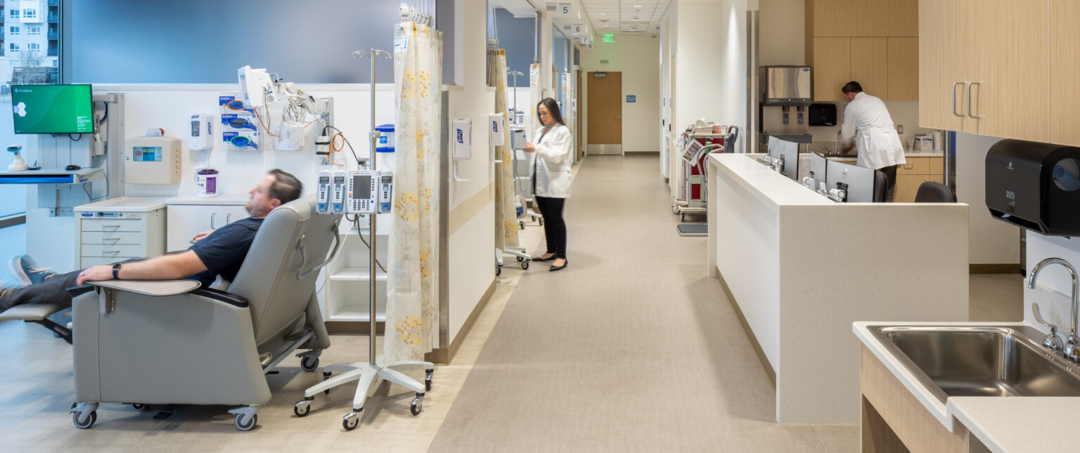 Hospital infusion center with a patient in a grey chair with medical equipment on the back wall looking down a hallway