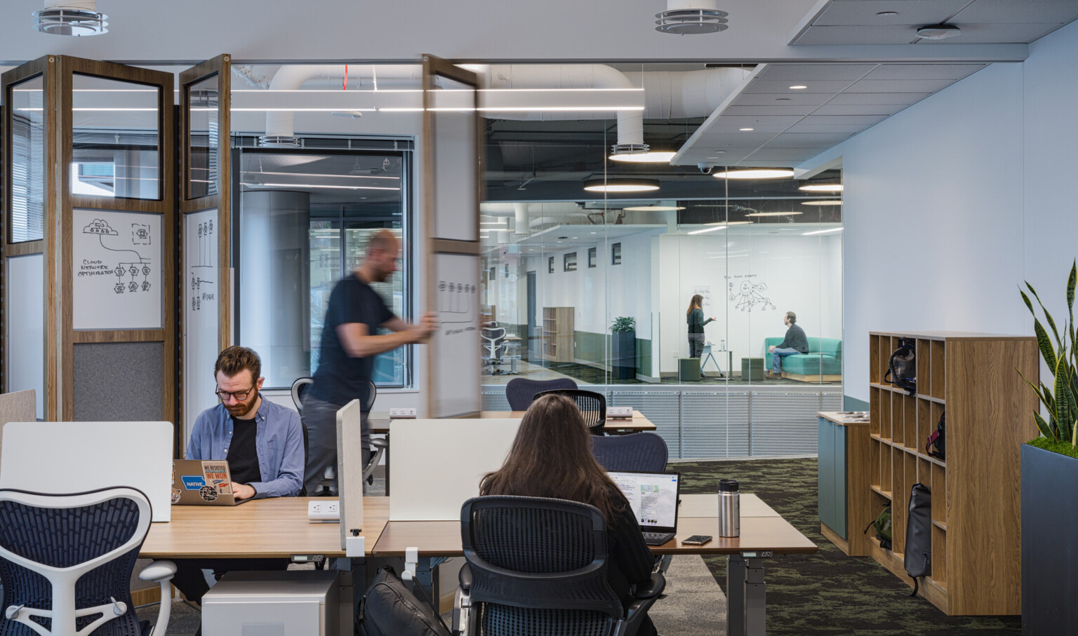 Open office workstations beside a folding partition featuring clear glass at the top and integrated whiteboard and pin-up panel