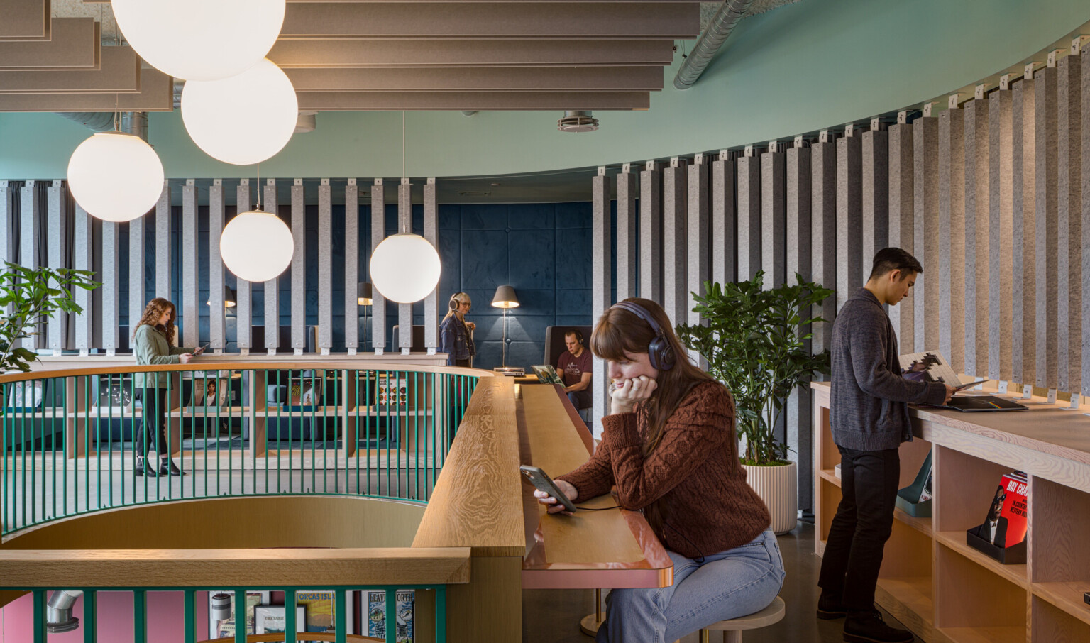 a woman on her phone with headphones sits at a radial wooden counter overlooking a mezzanine finished in wooden slats a woman on her phone with headphones sits at a radial wooden counter overlooking a mezzanine finished in wooden slats