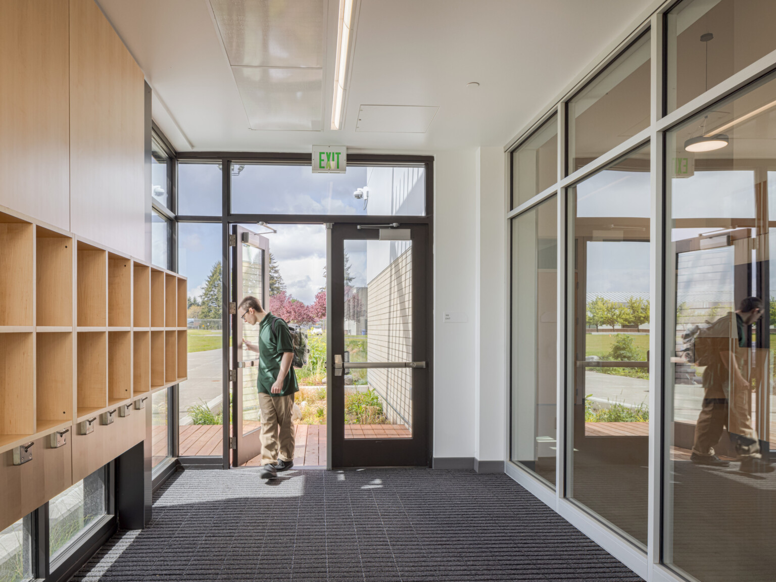 Looking down a hallway lined with glass windows on the right and shelves on the left at a door with a man entering from outside