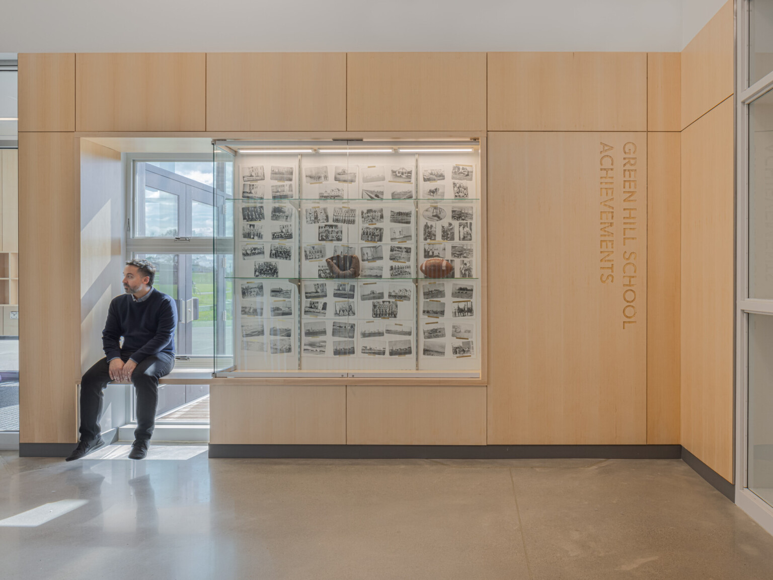 Wall with wooden accents with glass case filled with pictures and a baseball glove and football. Gentlemen sitting in a ledge next to the glass case