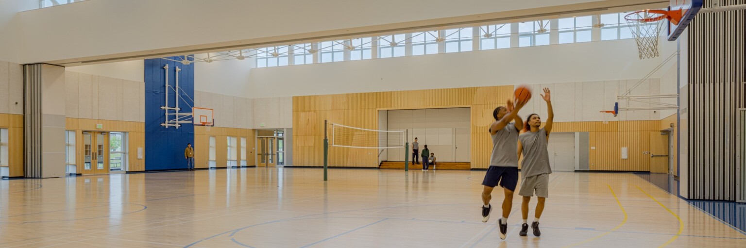 two men playing basketball at a community gym; skylights bring in natural light, light wood floor