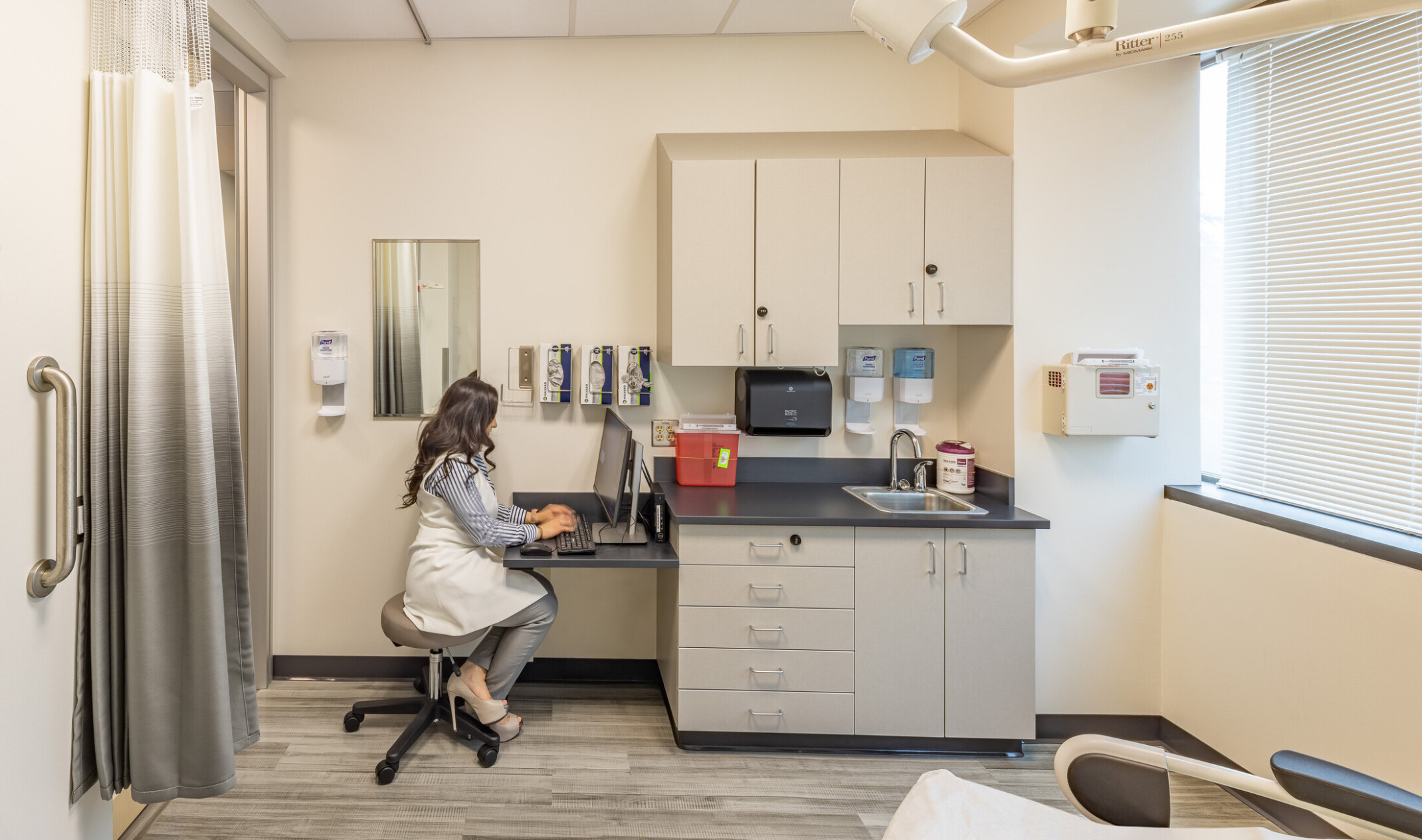 doctor working at a computer in an exam room; counter and cabinets next to her desk