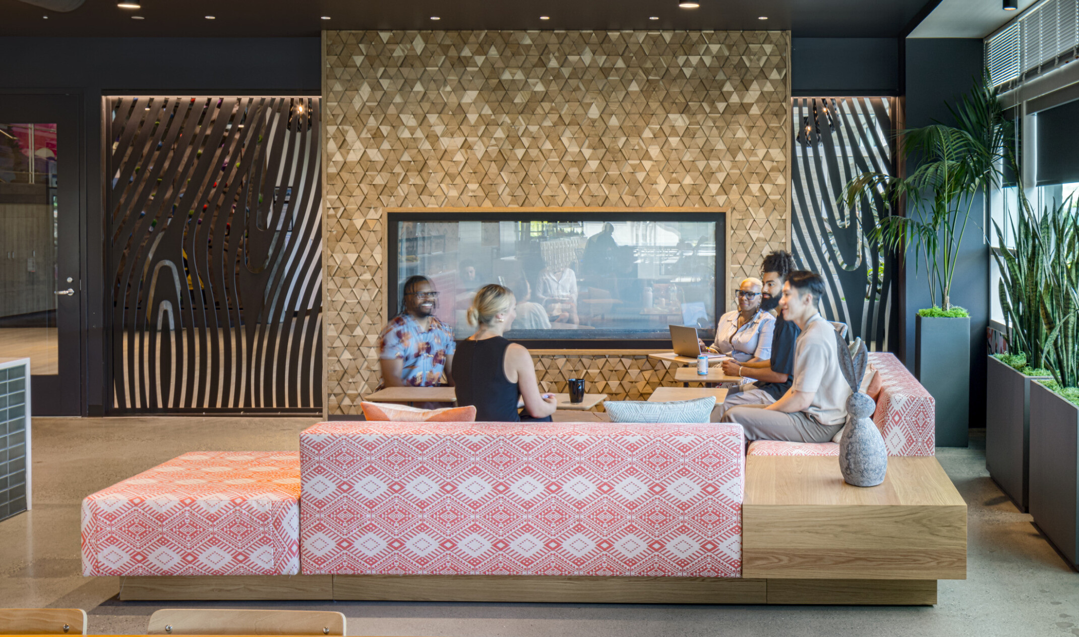workplace lounge with a group of people gathered in front of a modern gold tiled fireplace. Modern furnishings with red and white print, light wood accent pieces, black walls