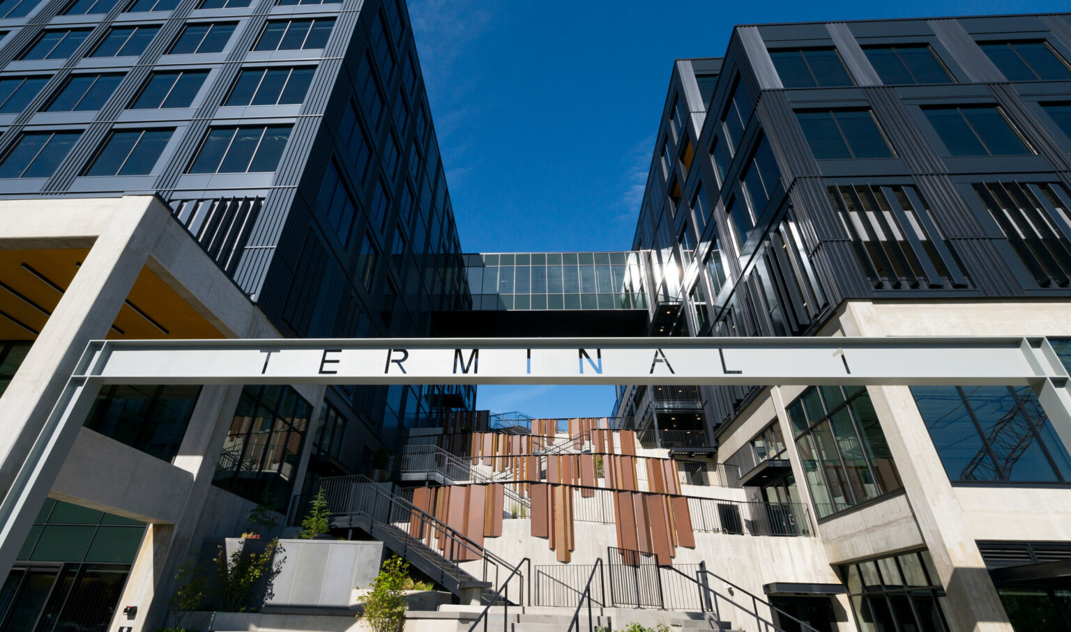Two modern buildings flanking a central staircase. An enclosed glass walkway bridges their upper floors. A sign with large letters spelling "TERMINAL" hangs prominently across the middle of the image
