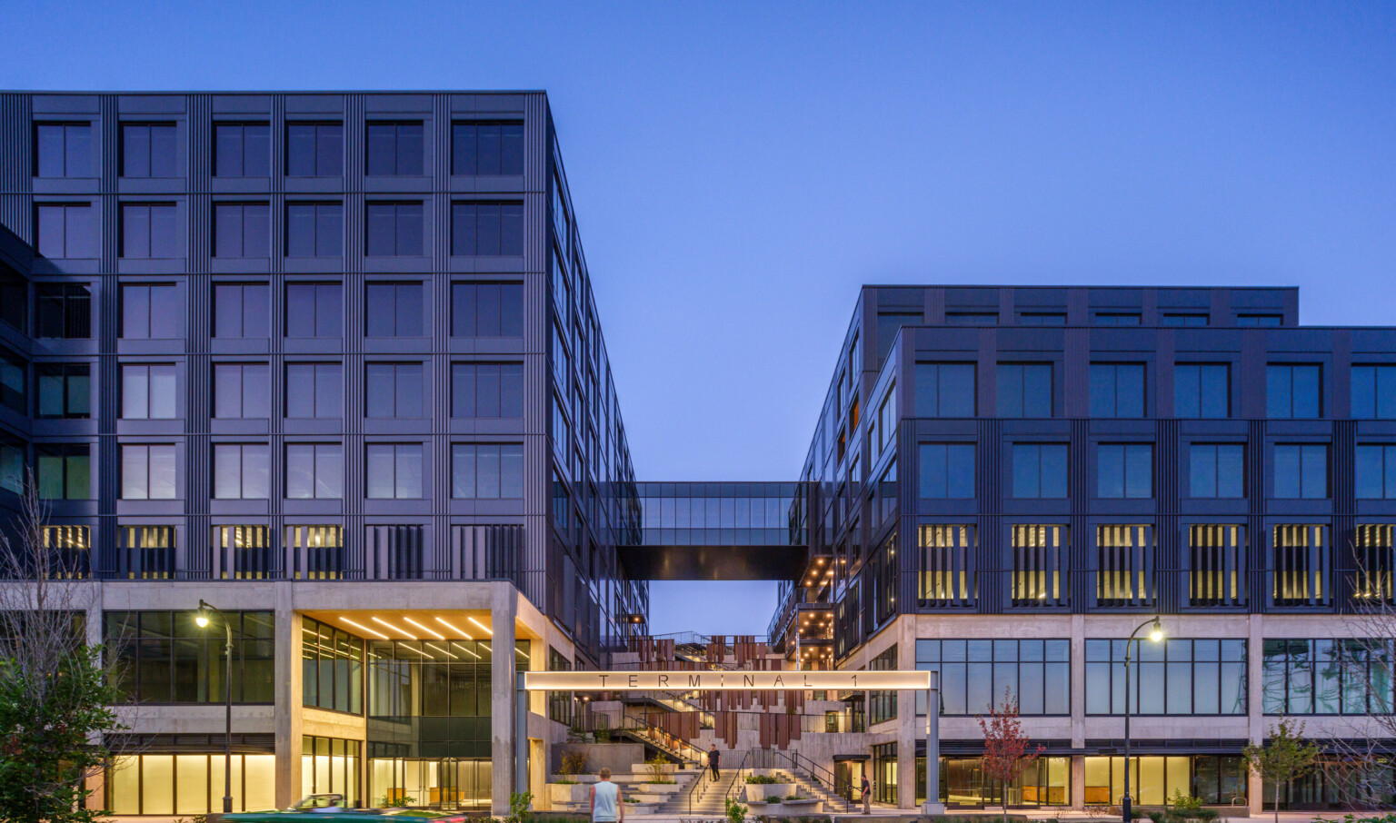 night view of multiple story mixed-use development, two large buildings connected by skyway, elevated bridgeway