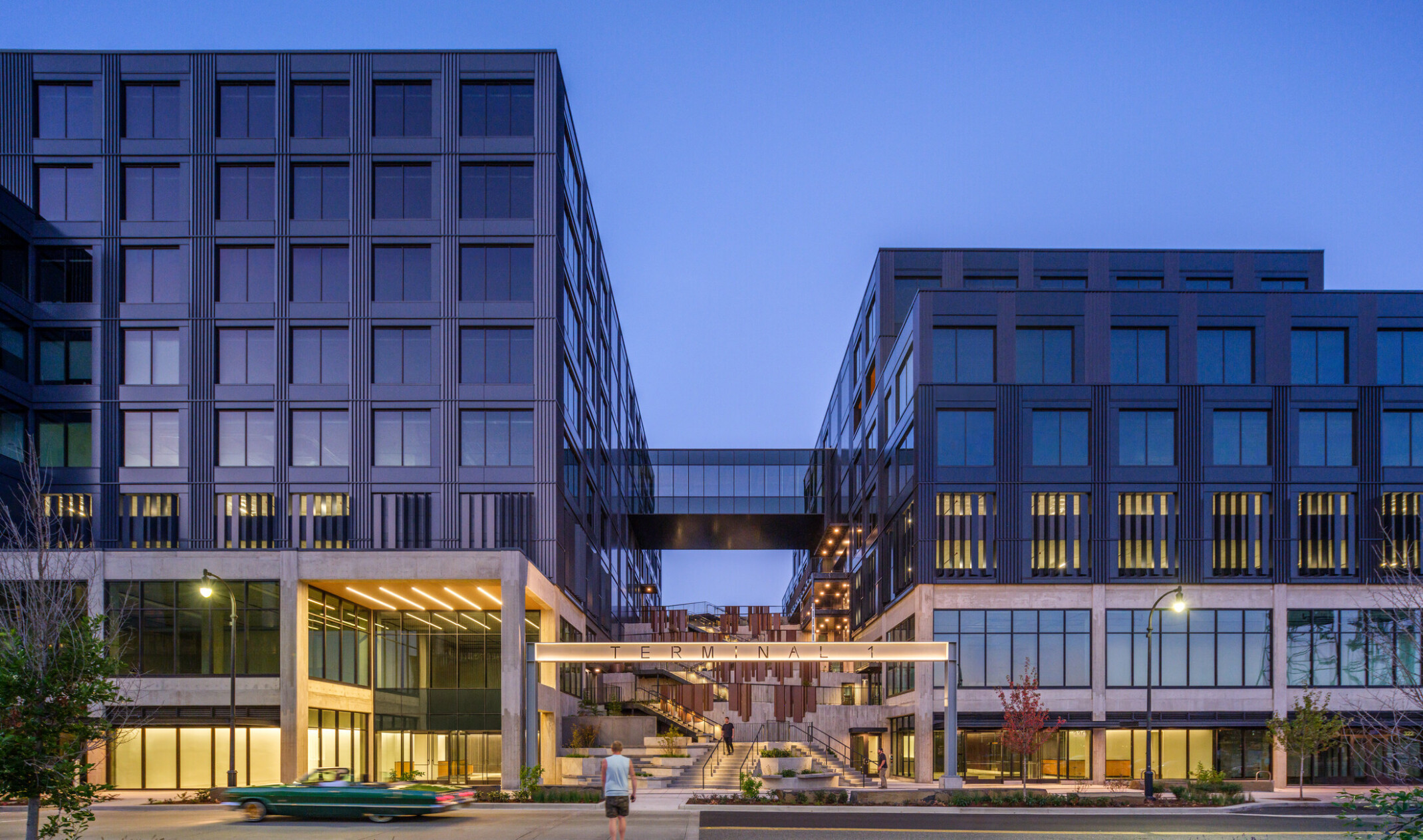 night view of multiple story mixed-use development, two large buildings connected by skyway, elevated bridgeway