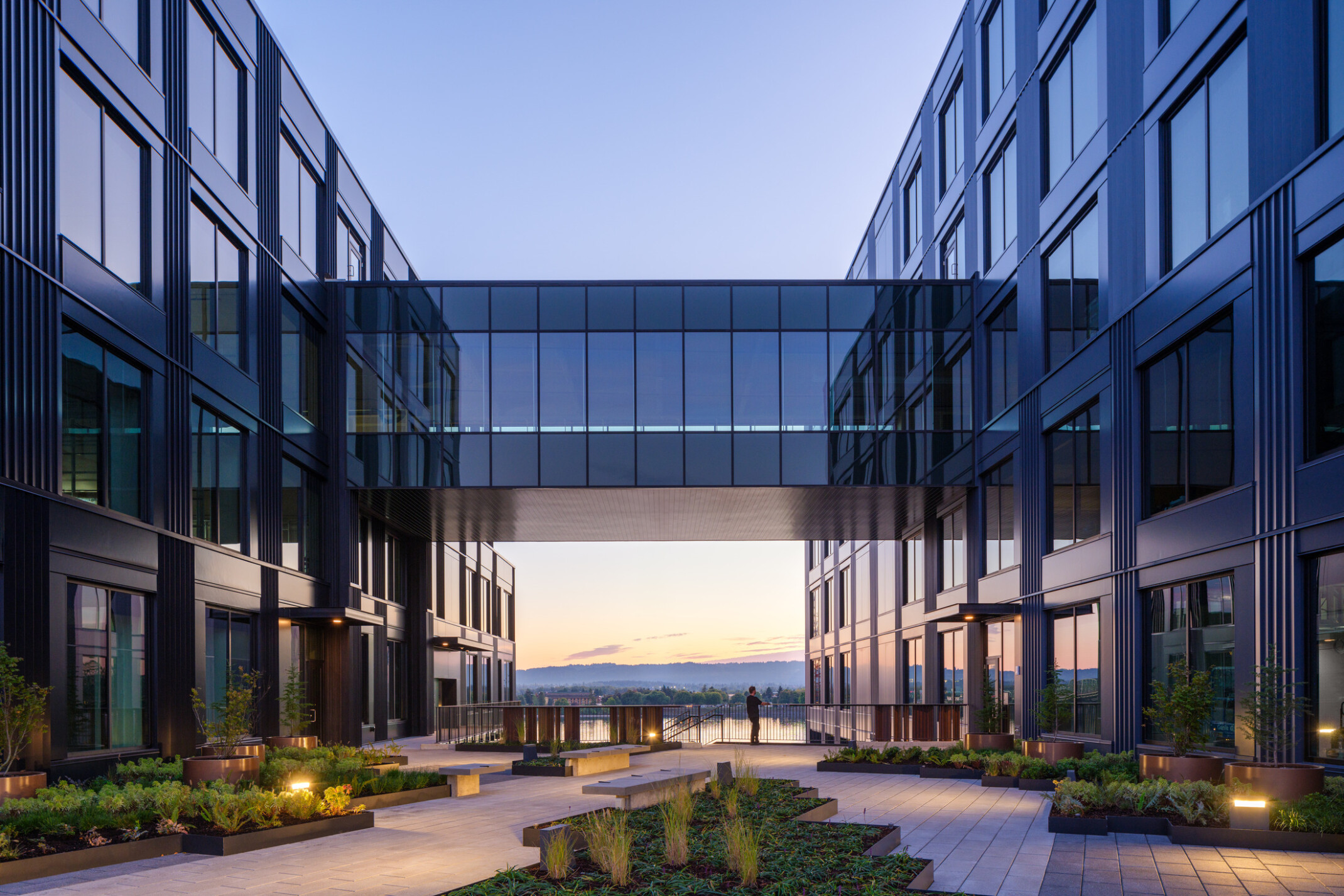 night view of development on the water in vancouver, wa; mixed-use development with dark facade, elevated walkway, skyway connecting two buildings, courtyard for wellness