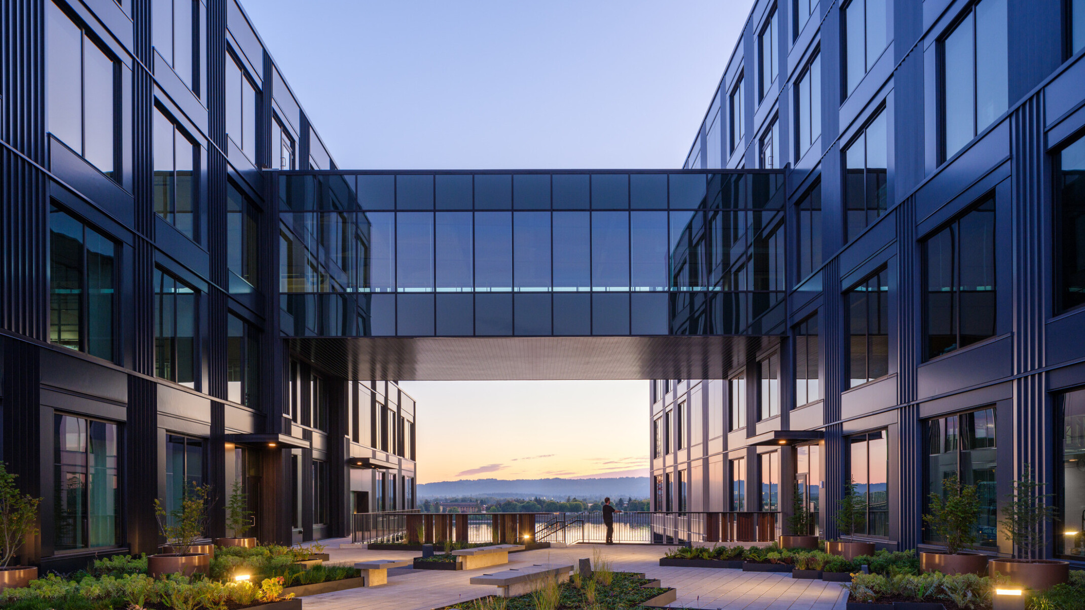 night view of development on the water in vancouver, wa; mixed-use development with dark facade, elevated walkway, skyway connecting two buildings, courtyard for wellness