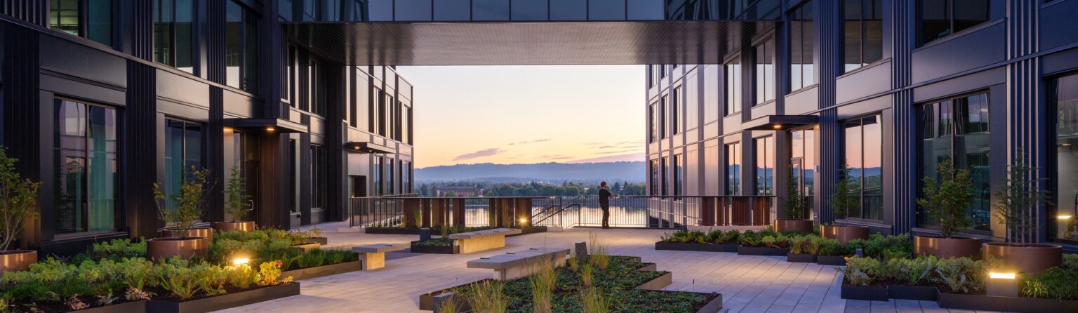 night view of development on the water in vancouver, wa; mixed-use development with dark facade, elevated walkway, skyway connecting two buildings, courtyard for wellness