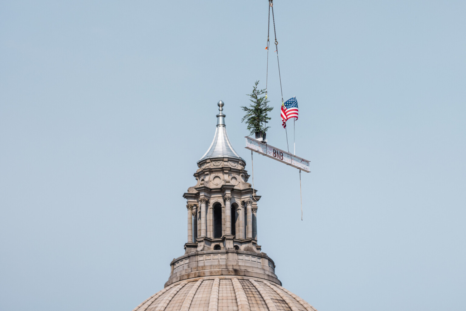 Helicopter flies steel beam bearing a potted cedar tree and the US flag past the Washington State Capitol dome on its way to the Pritchard Building. Photo Courtesy of Taras Yaremchuk/BNBuilders Helicopter flies steel beam bearing a potted cedar tree and the US flag past the Washington State Capitol dome on its way to the Pritchard Building. Photo Courtesy of Taras Yaremchuk/BNBuilders