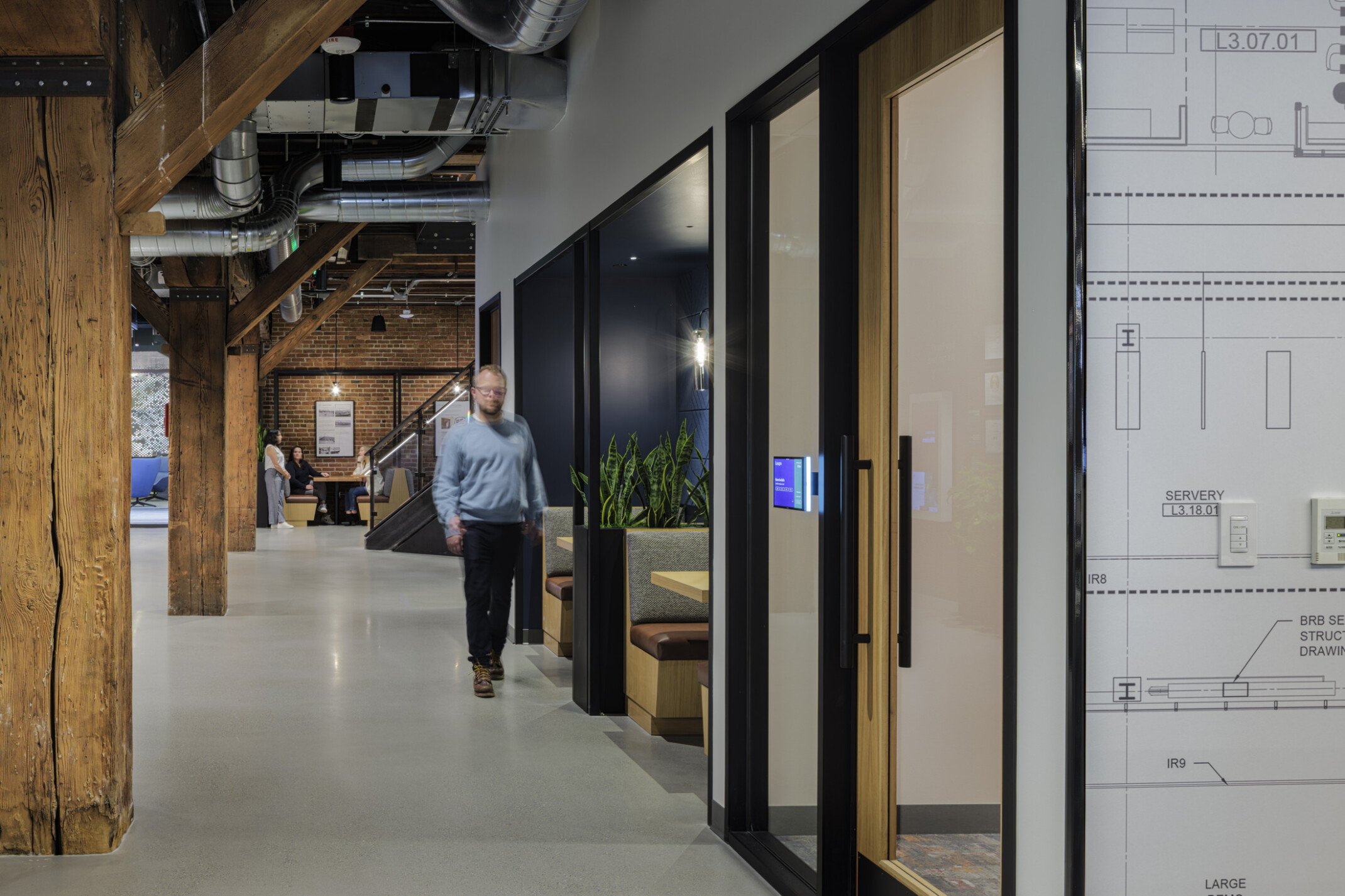 Corridor lined with original timber columns at BNBuilders' Seattle headquarters, passing small meeting rooms and open banquette seating, with a brick wall and staircase beyond.