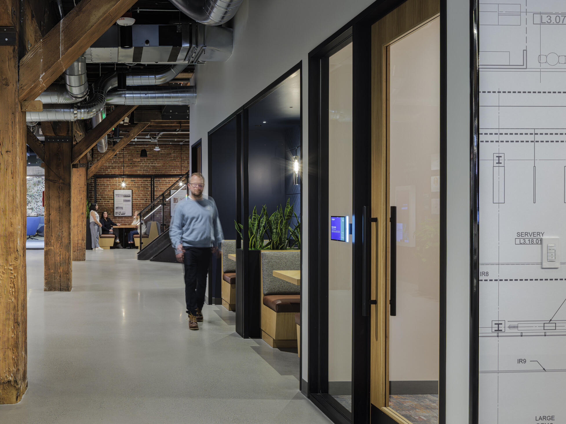 Corridor lined with original timber columns at BNBuilders' Seattle headquarters, passing small meeting rooms and open banquette seating, with a brick wall and staircase beyond.