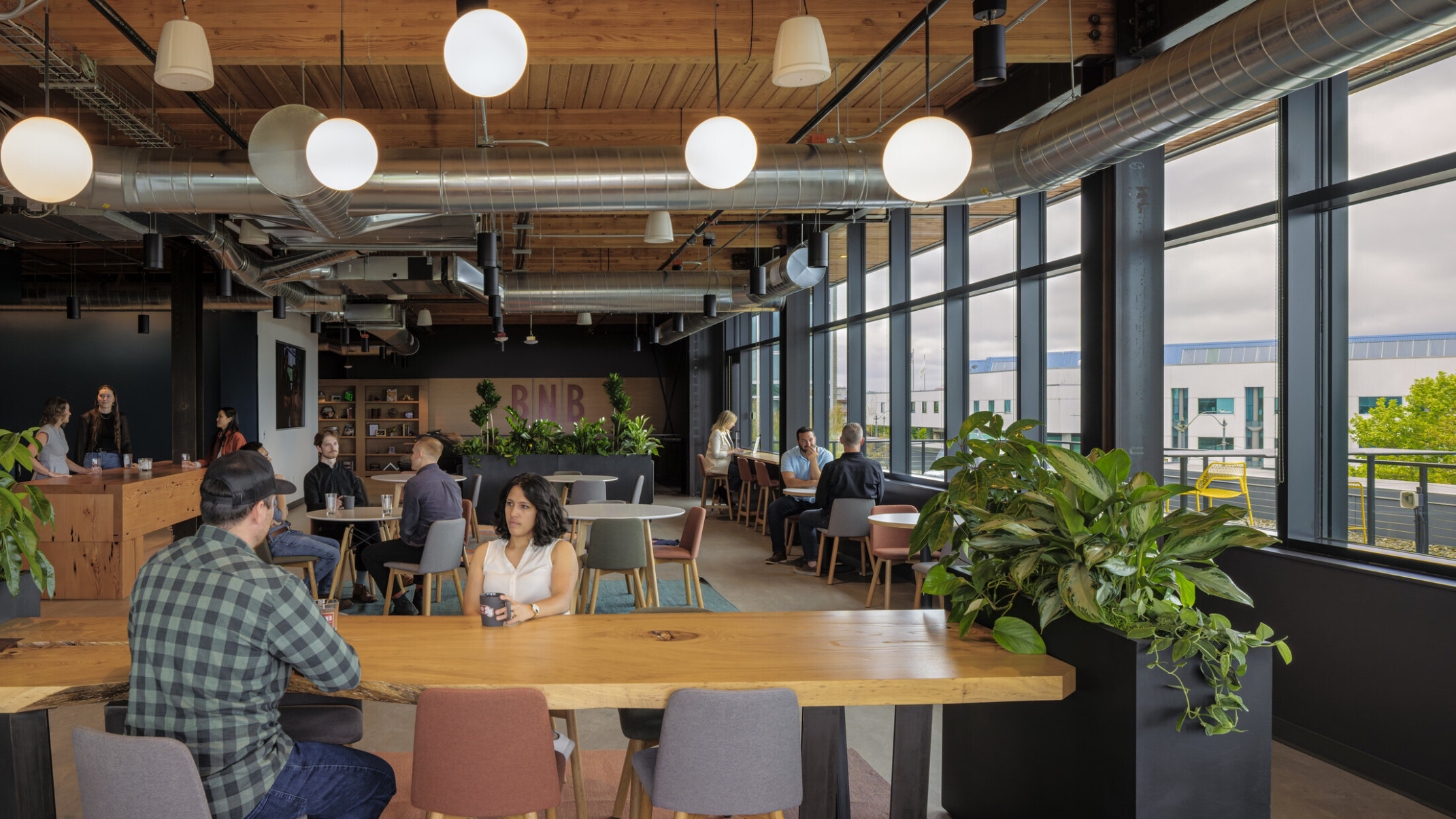 Employees gather in a work café with large windows with views outside; mixing casual conversations with heads-down work